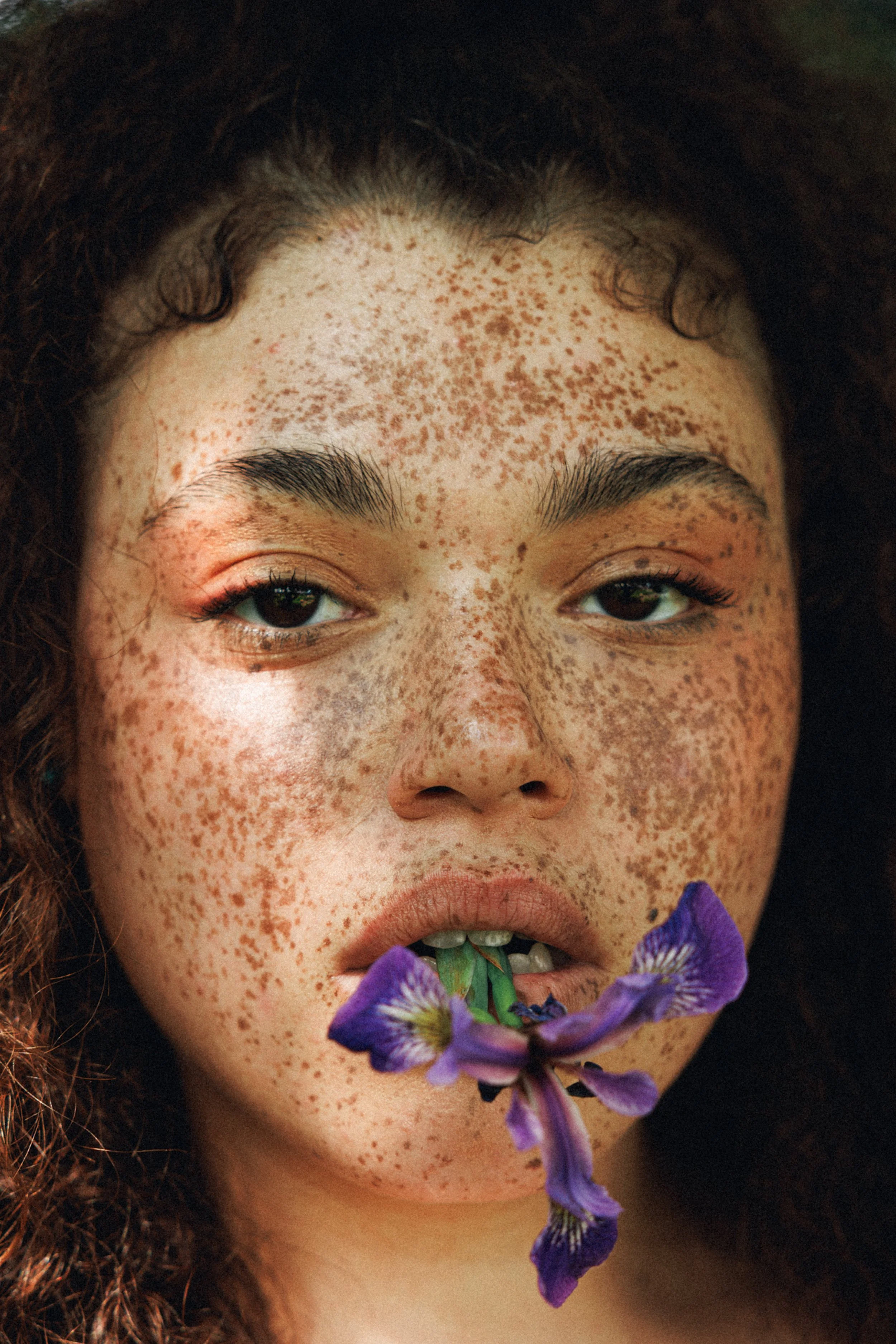 Close-up of a person's face with freckles, slightly parted lips holding a purple flower, dark curly hair, and brown eyes.