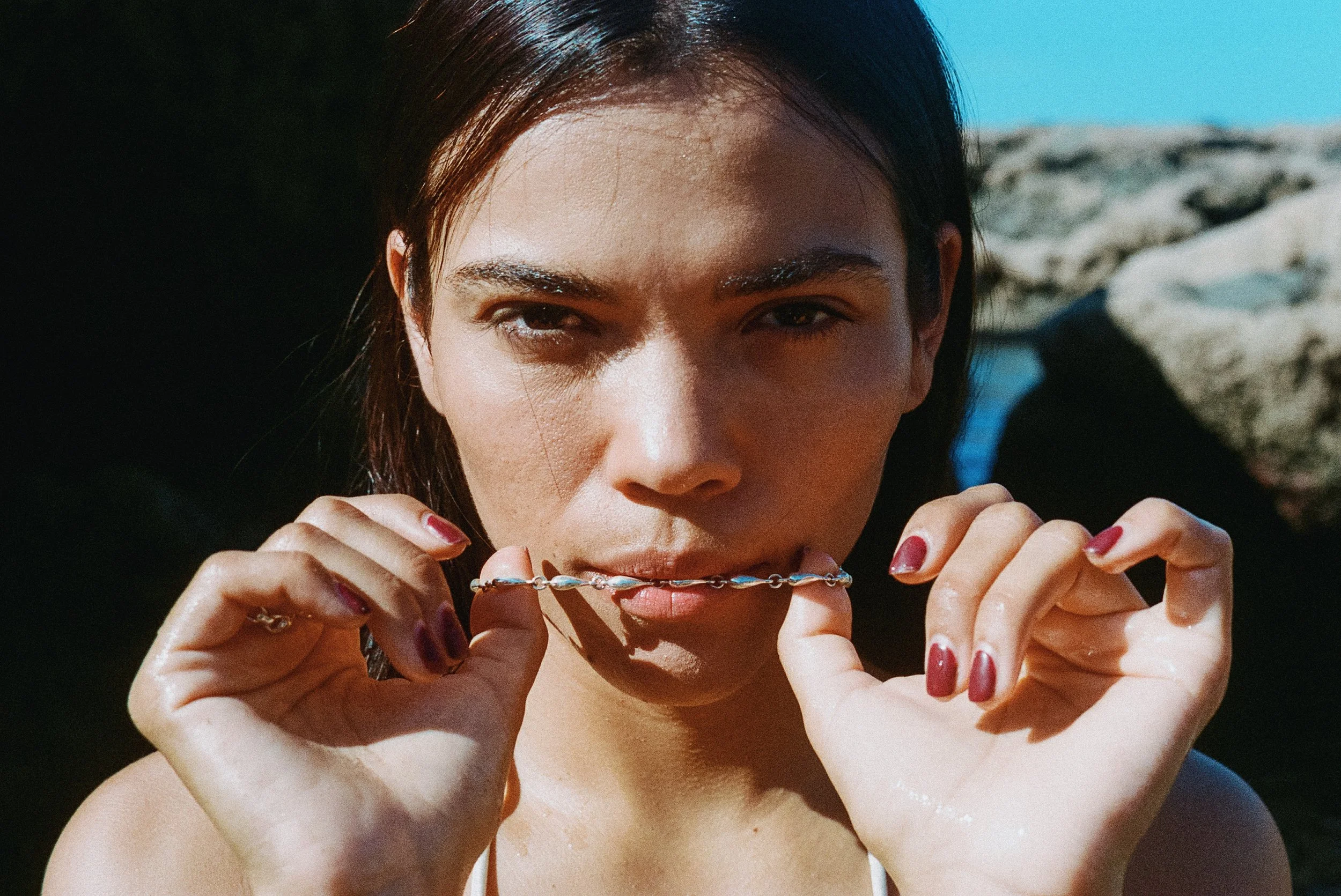 A young woman with dark hair is holding a silver necklace with blue and white stones close to her face on a beach with rocks and ocean in the background.