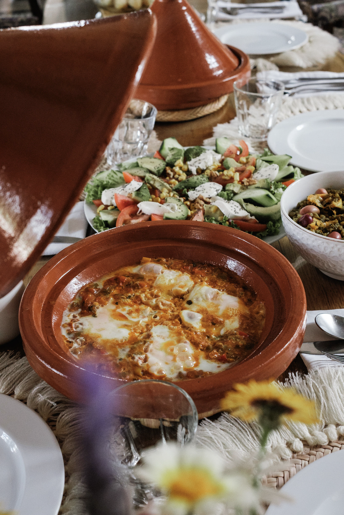 A table set with a variety of Mexican dishes, including an individual claypot of baked eggs with cheese, a mixed salad with tomatoes, cucumbers, corn, and cheese, and a bowl of beans with vegetables. The table is decorated with a woven tablecloth, plates, glasses, and silverware.