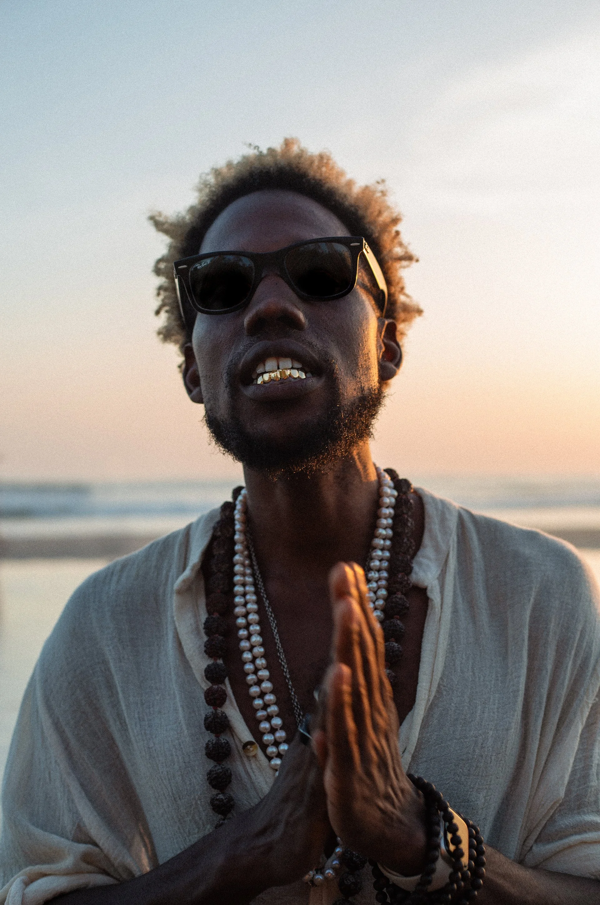 A man with dark skin, curly hair, wearing sunglasses, gold teeth, and layered necklaces, is standing outdoors during sunset, with a beach and ocean in the background. He is dressed in a light-colored, loose shirt and appears to be holding something in his hand.