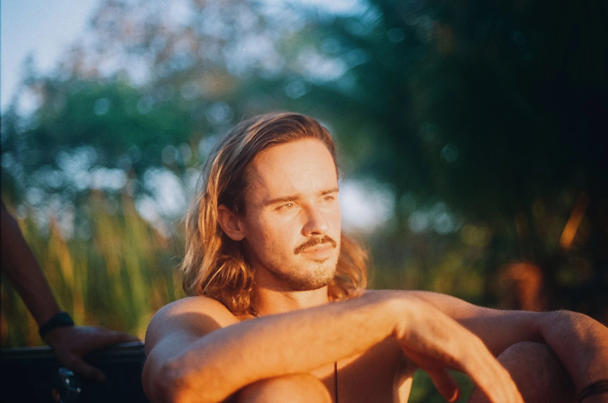 A man with long hair and a beard outdoors, with greenery and blurred light in the background, appears to be doing yoga or meditation.