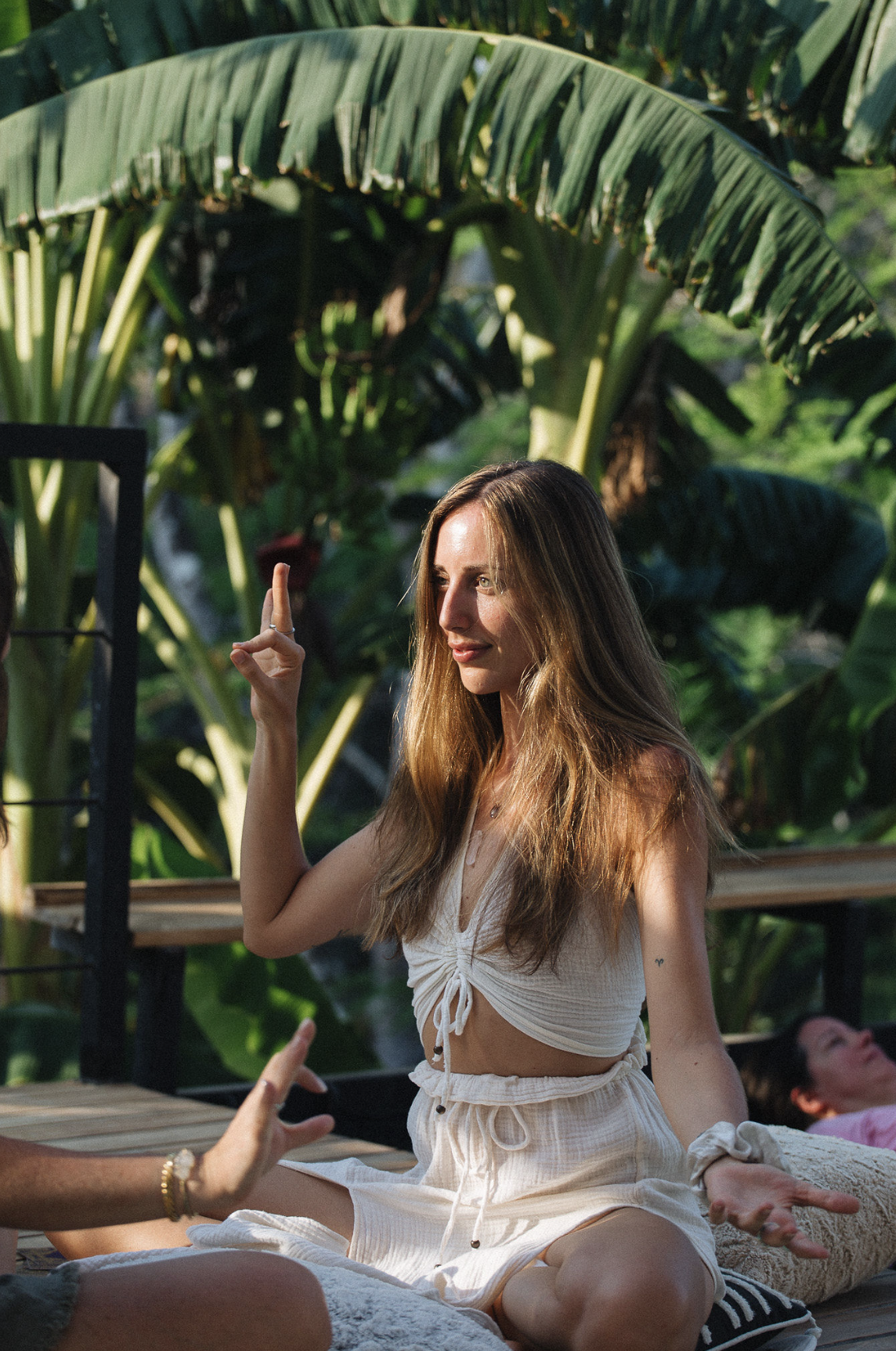 A young woman sitting cross-legged outdoors, making a hand gesture, with large green banana leaves in the background, enjoying a sunny day.