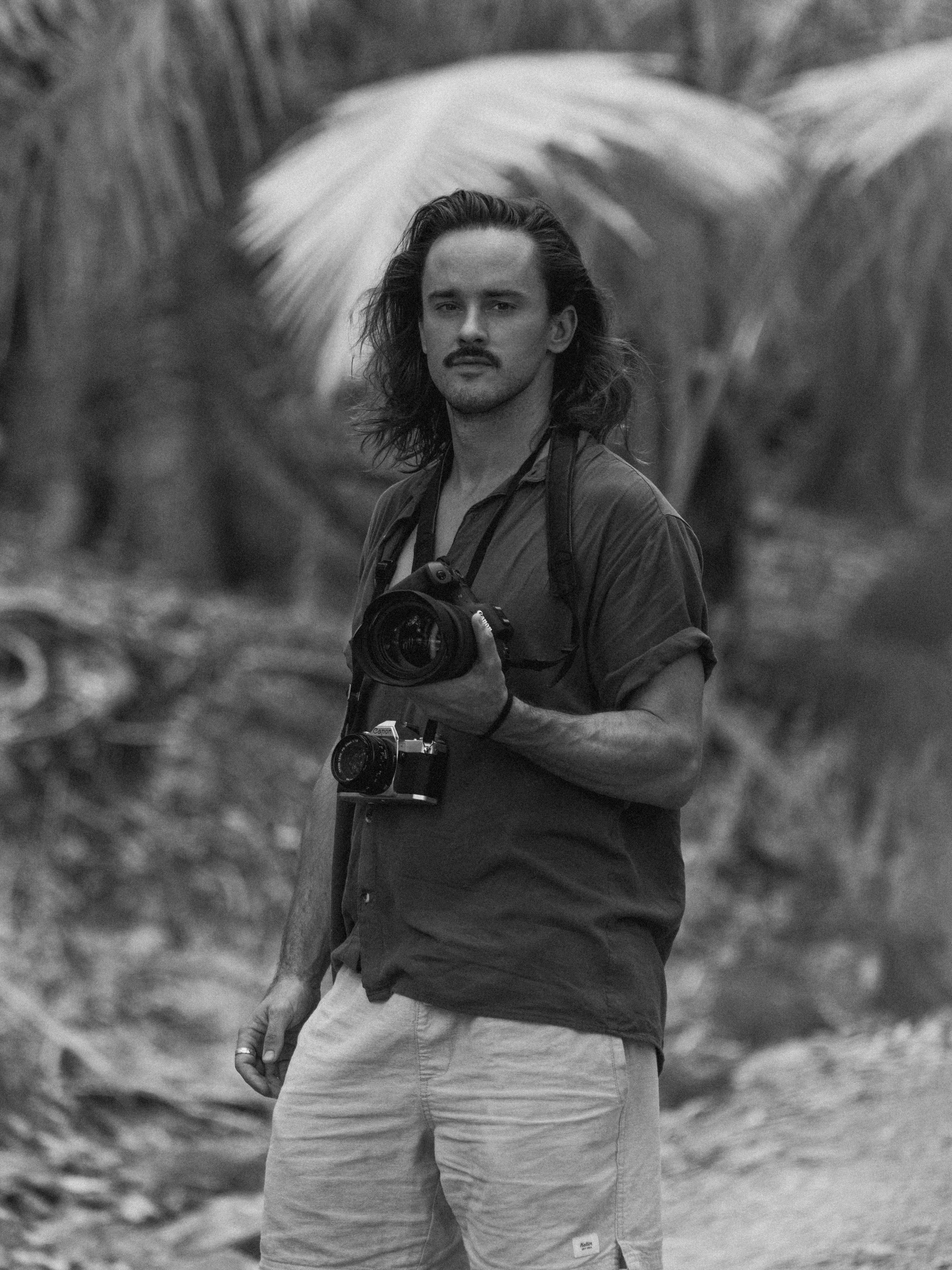 A man with long hair and a mustache taking a selfie outdoors with a camera hanging around his neck.