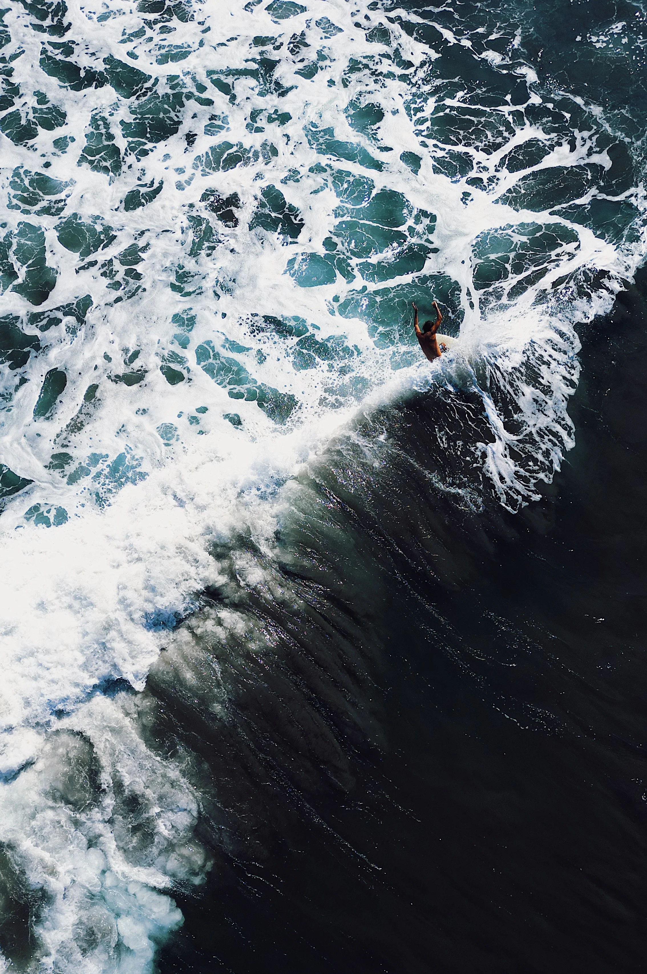 An aerial view of the ocean showing white foamy waves and a person swimming or surfing in the water.