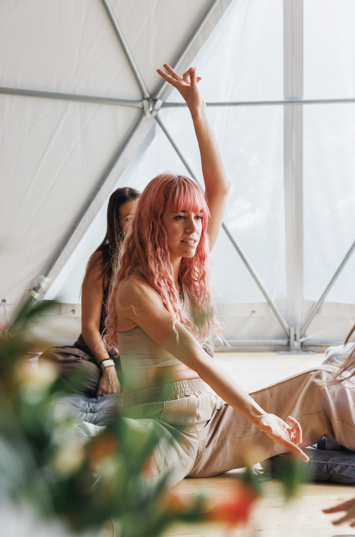 A young woman with pink hair is sitting on the floor in a modern building with large windows, raising her hand during a discussion or class.
