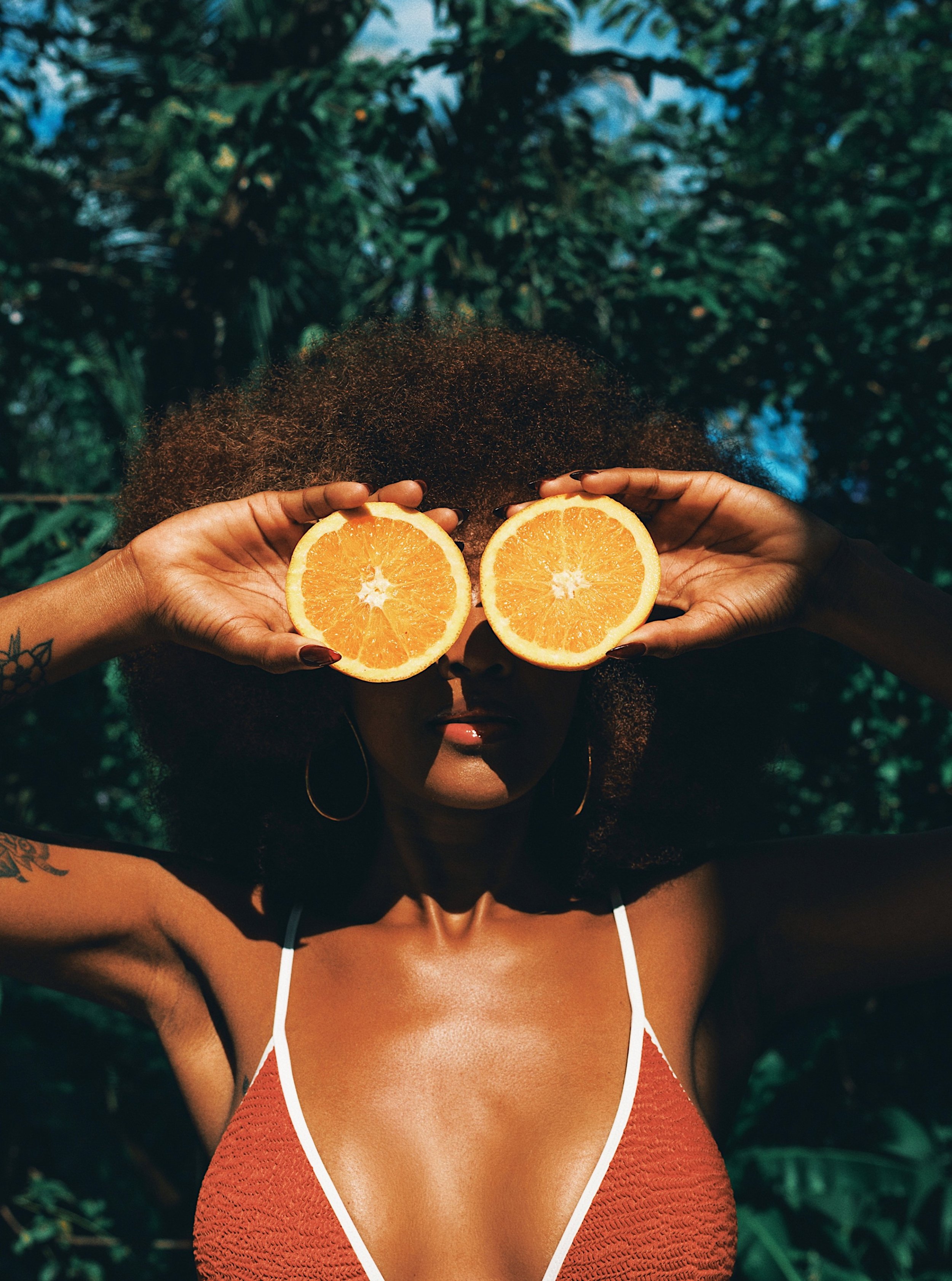 A woman with curly hair holds two halves of an orange in front of her eyes. She is wearing a red sleeveless top with white trim and has tattoos on her arms. The background is lush green foliage.