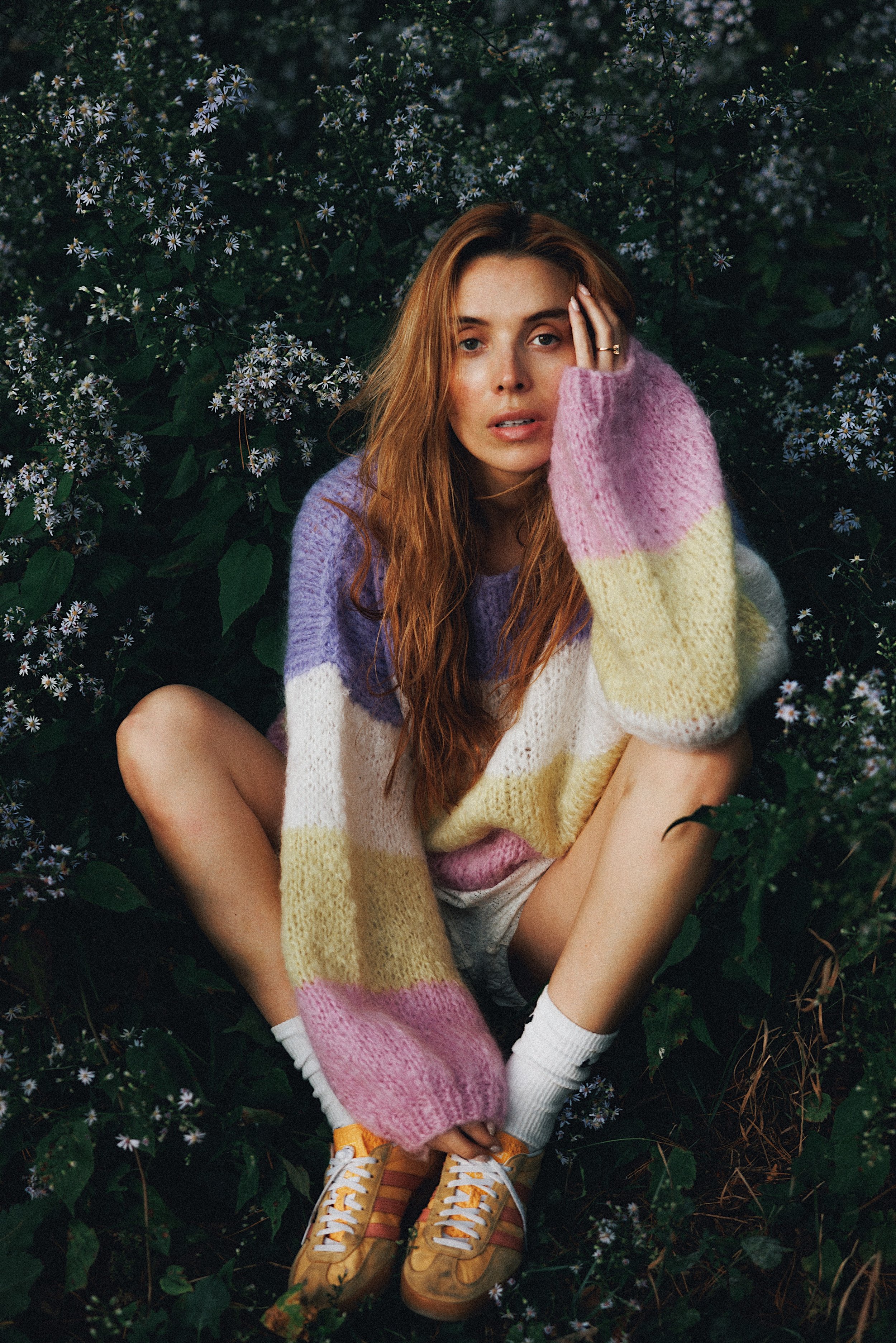 A woman with long red hair and light skin sitting among greenery and small white flowers, wearing a pastel-colored oversized sweater, white socks, and yellow sneakers.