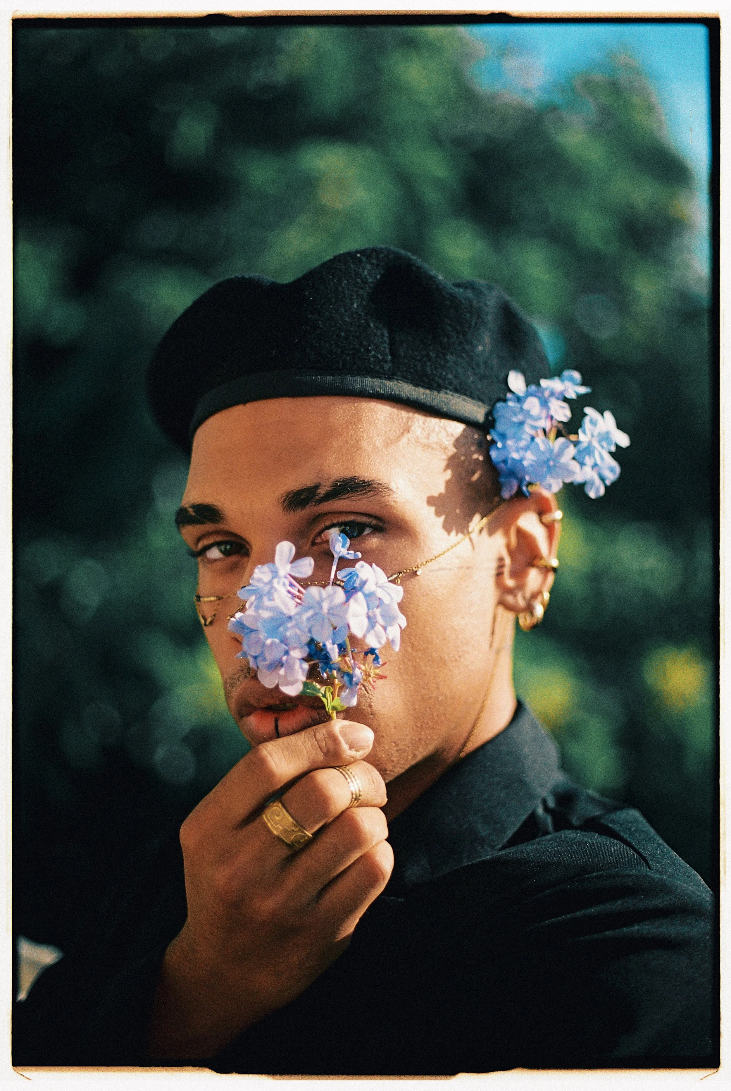 A person with a dark beret, multiple gold earrings, and rings holding a bunch of blue flowers, looking at the camera with a blurred green background.