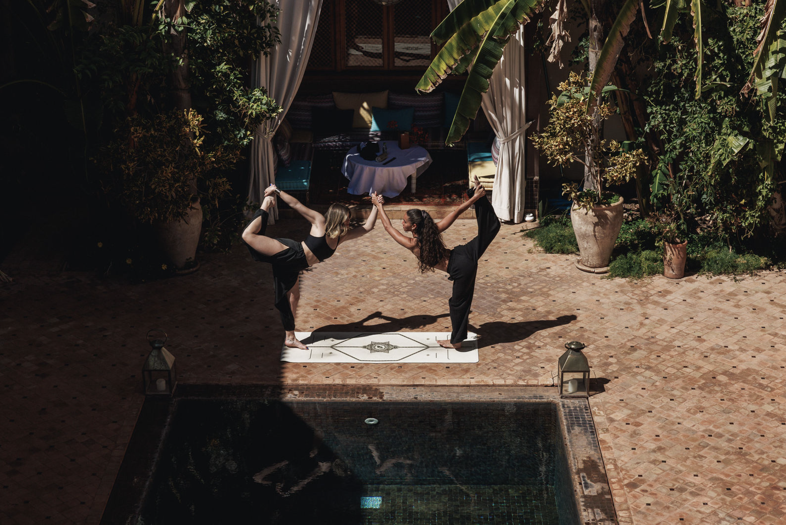 Two women practicing acro yoga outdoors on a brick patio surrounded by potted plants, with one woman balancing on her foot on a mat while the other woman supports her by holding her foot and hand, both facing each other.
