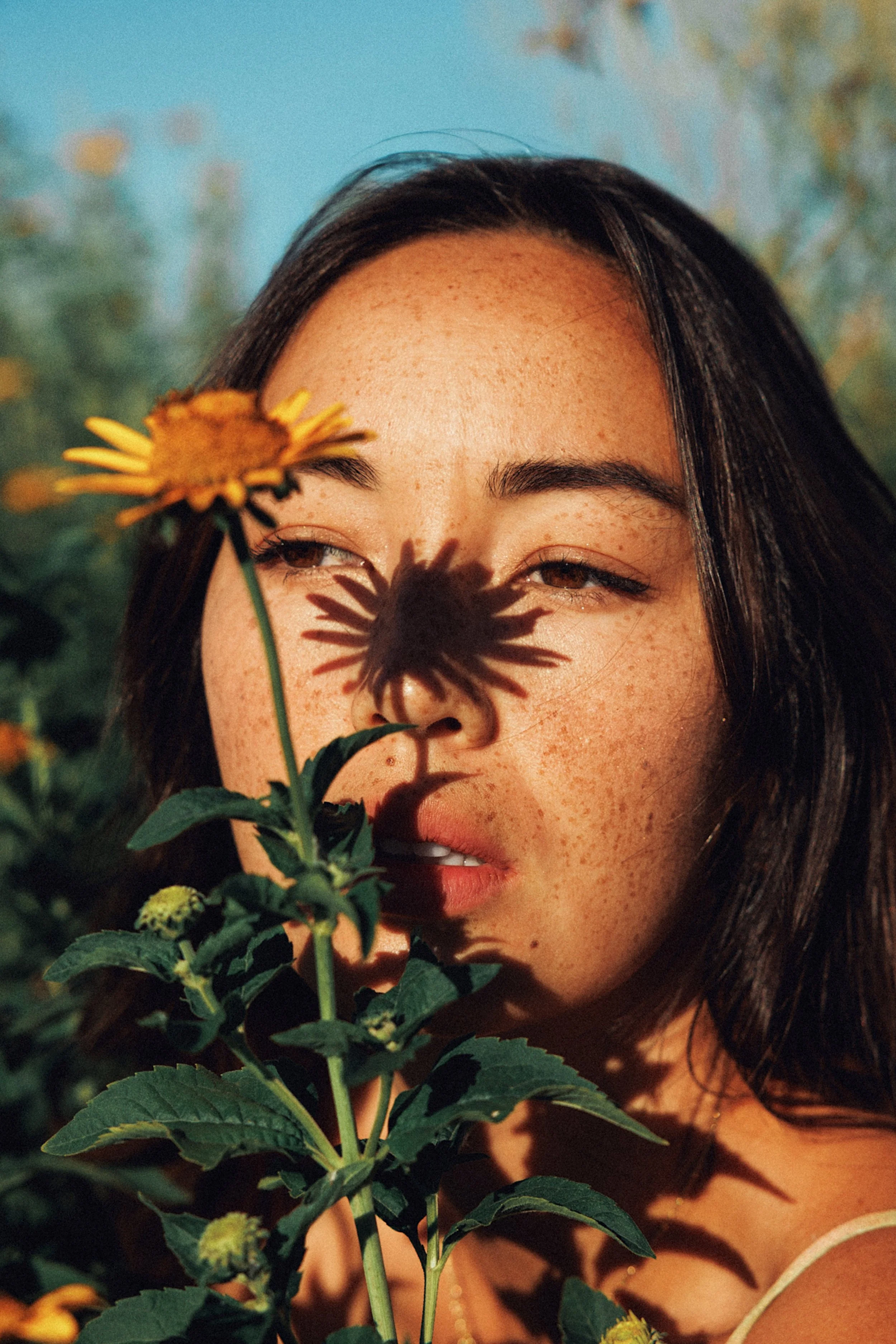 A woman with dark hair and freckled skin holding a yellow flower, casting a shadow on her face, outdoors with greenery and blue sky in background.