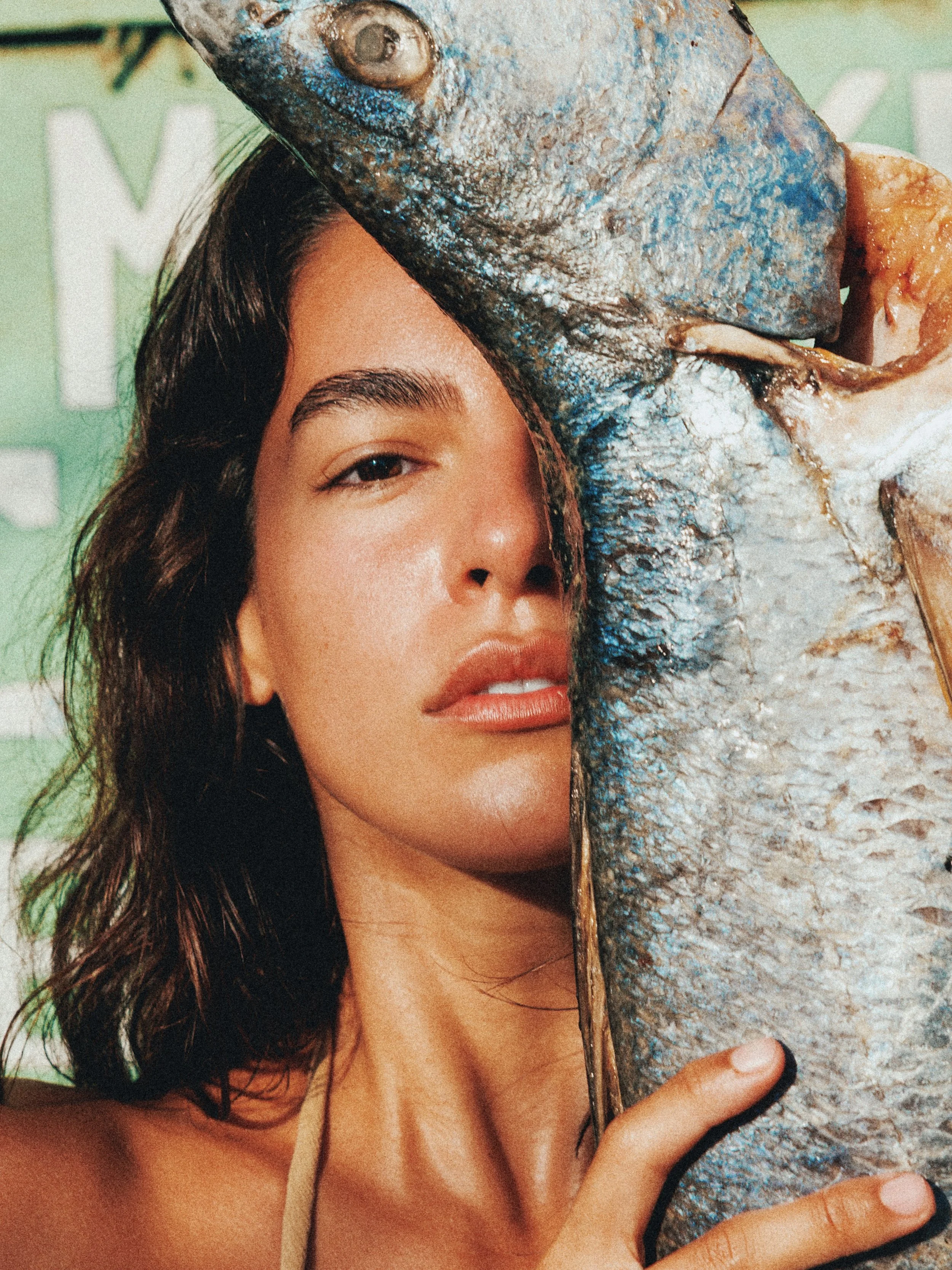 A woman with dark brown hair and natural makeup holds a large fish close to her face, partially covering it. The woman is smiling slightly, and the background is green with some white text.