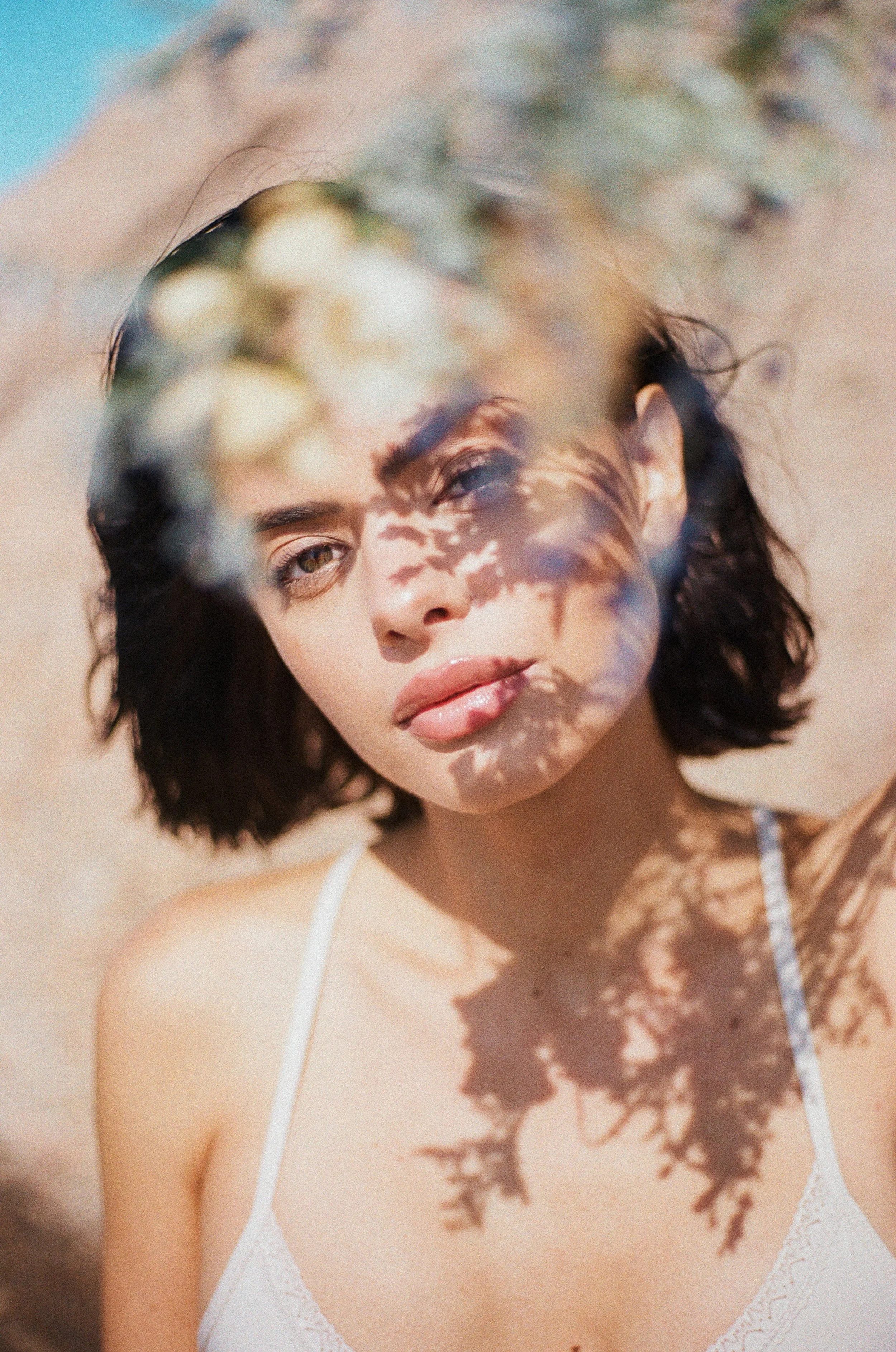 A woman with short dark hair and light brown eyes sitting outdoors, wearing a white top. The shadow of tree leaves is cast on her face and upper chest, creating a pattern.