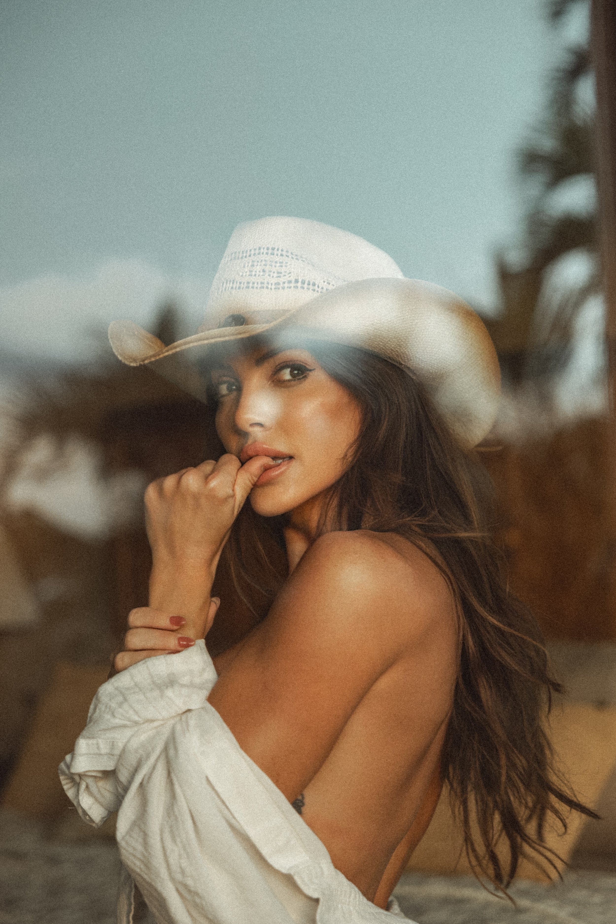 A woman with long wavy brown hair wearing a large straw cowboy hat and a white off-shoulder dress, standing outdoors with blurred trees in the background.