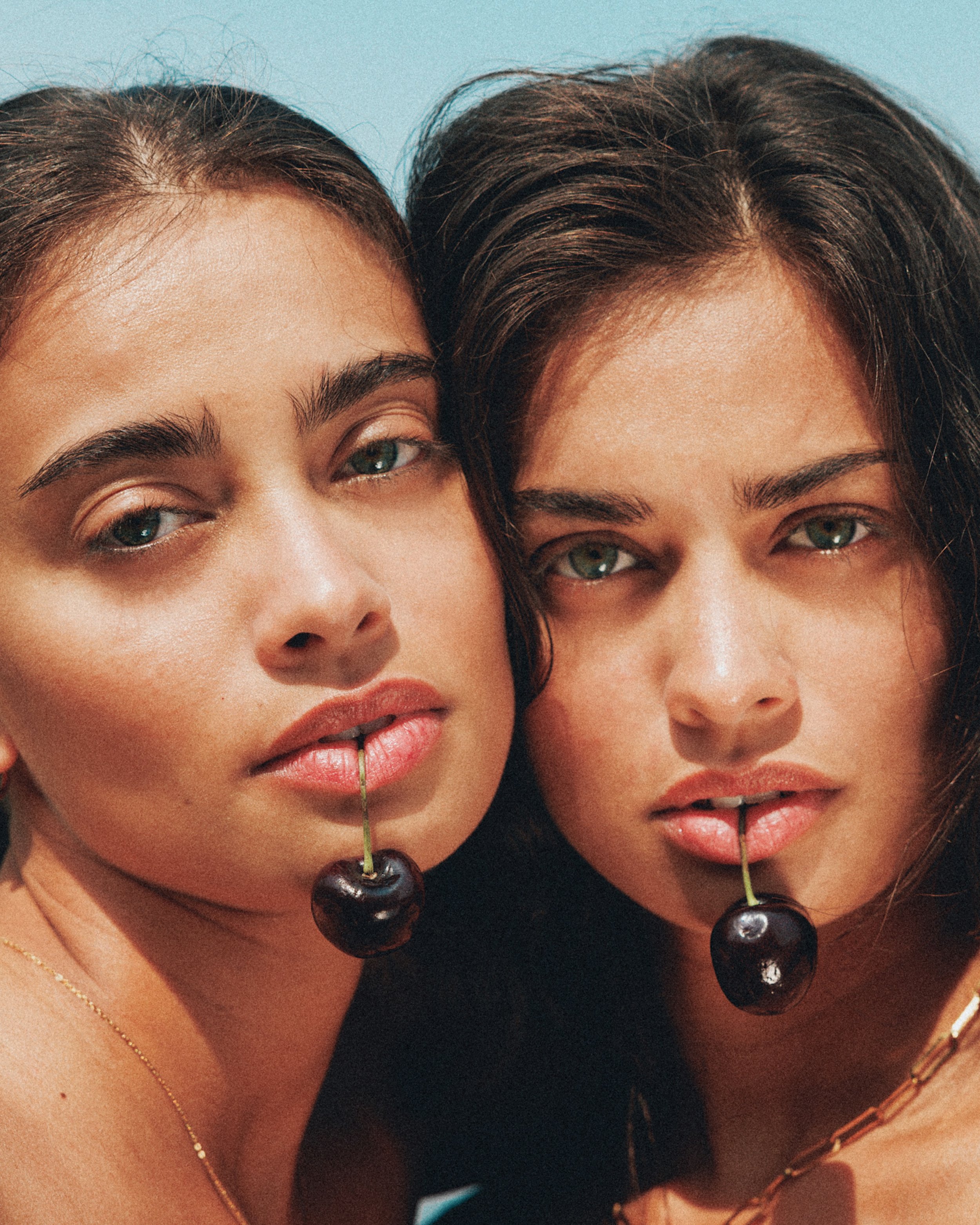 Close-up of two women with dark hair and green eyes, holding cherries with stems in their mouths.