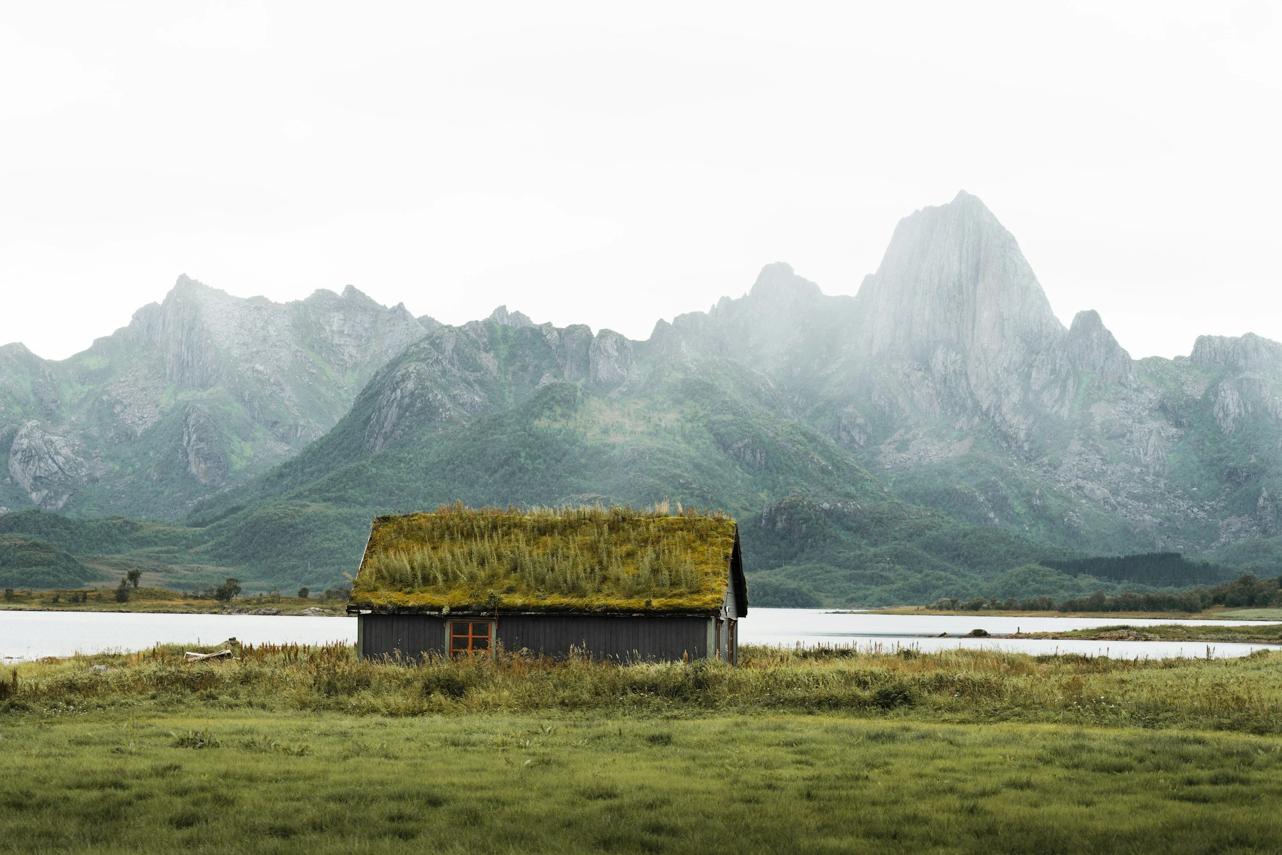 A small house with a grass-covered roof sits near a calm lake, with mountains in the background.