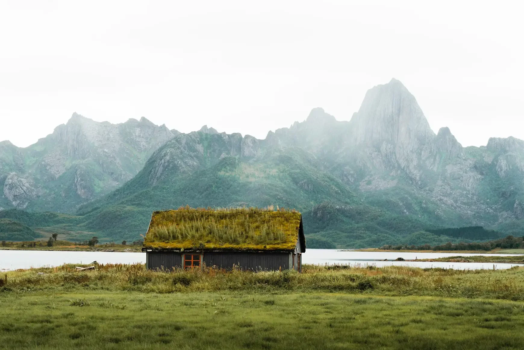 A house with a moss-covered roof sits on a grassy field near a calm body of water, with tall mountains in the background under an overcast sky.