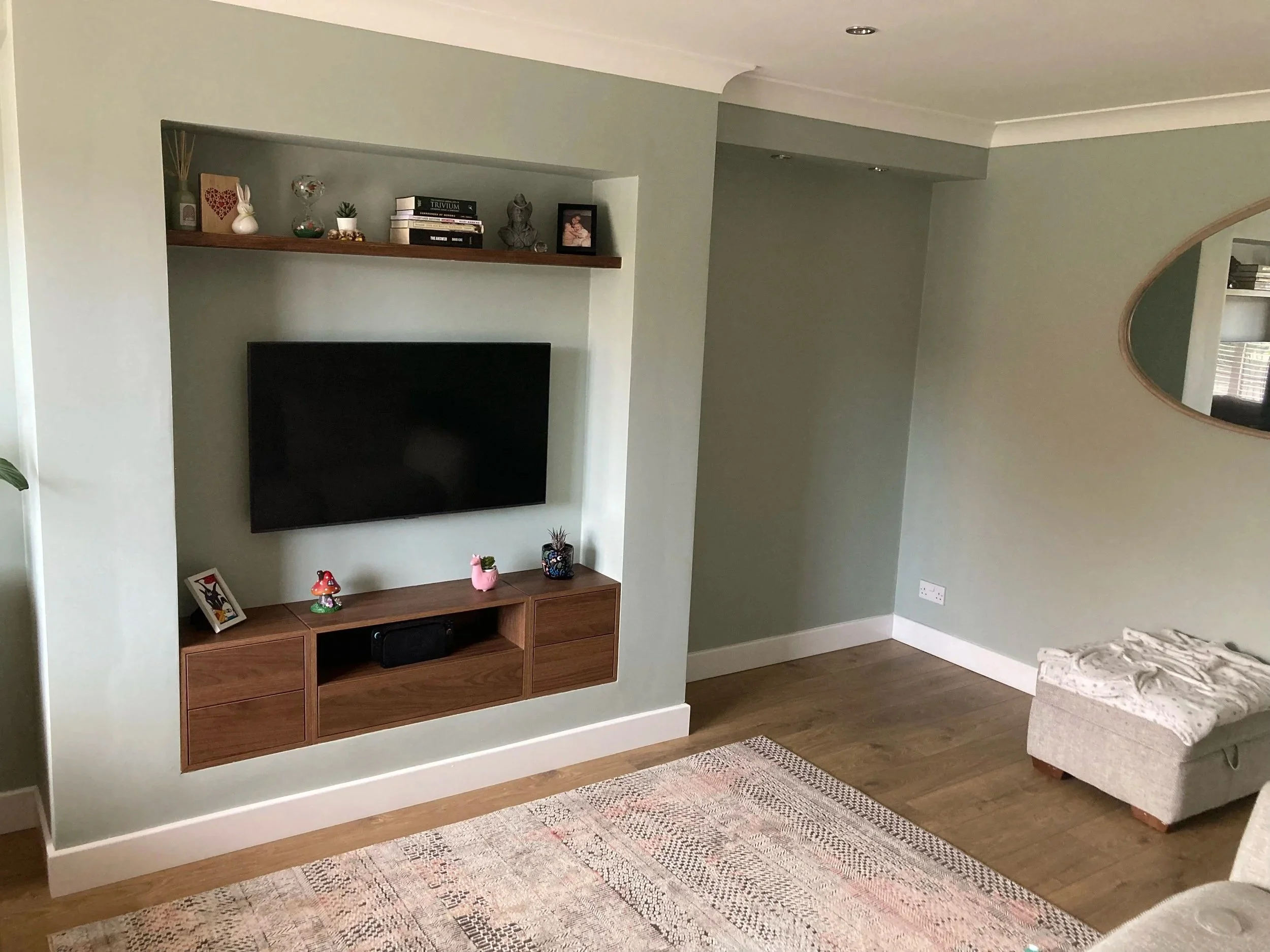 Living room wall with mounted flat-screen TV, wooden shelves decorated with small plants, books, and decorative items, beige and green walls, a patterned area rug, and part of a couch and a mirror visible.