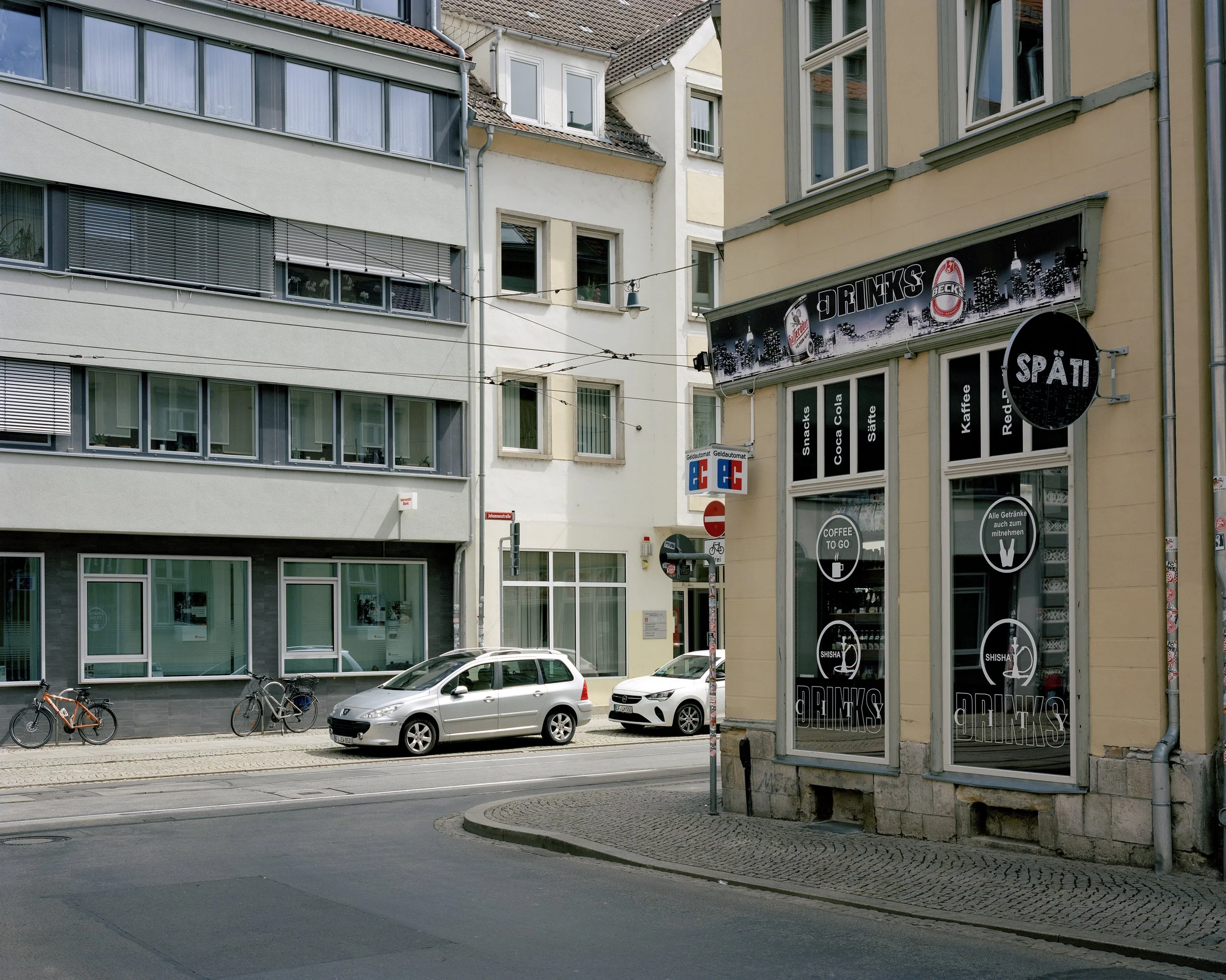 Ansicht von der Ecke Johannesstraße in Erfurt mit modernen und alten Gebäuden, zwei Fahrrädern, mehreren parkenden Autos, einer Werbung für den Späti, der an der Ecke Getränke verkauft. Im Hintergrund sind Straßenlaternen und die Straßenbahnlinie.