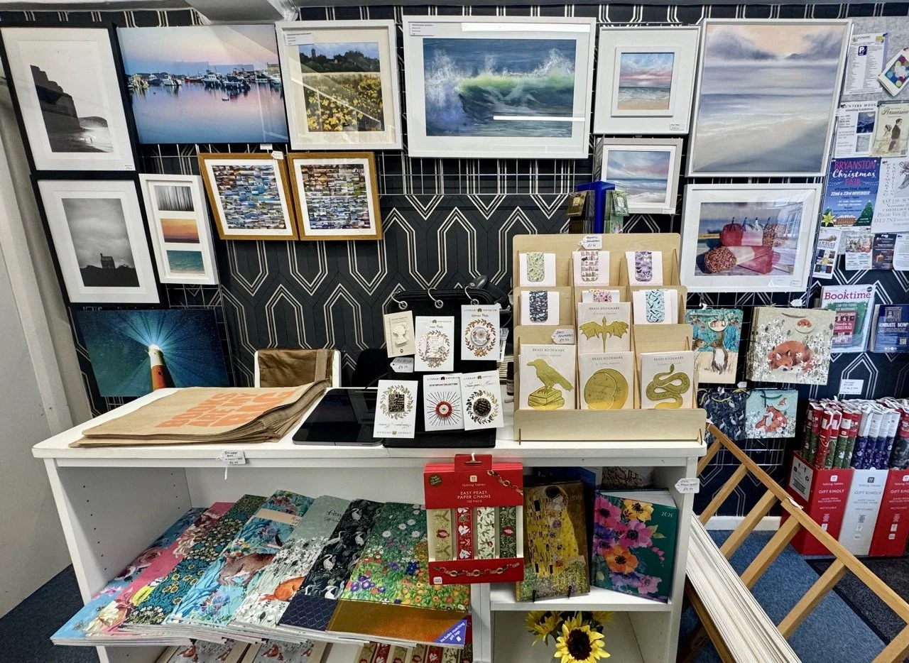 Art and gift shop display with framed landscape and seascape photographs on the wall, jewelry, greeting cards, and decorative notebooks on the table, and Christmas decorations in the background.