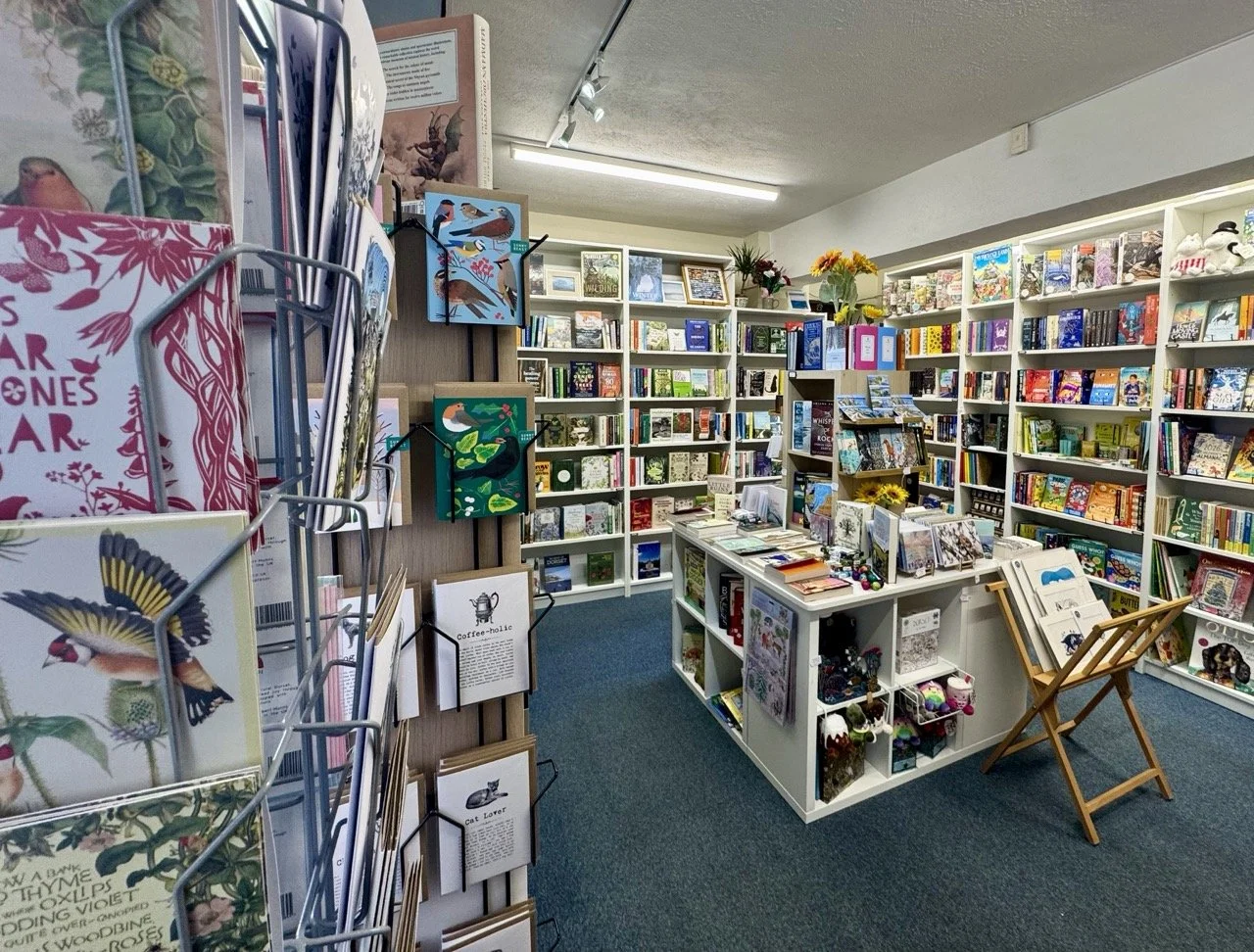 Interior of a bookstore with white shelves filled with books, a table with books and toys, and a display rack with greeting cards on the side.