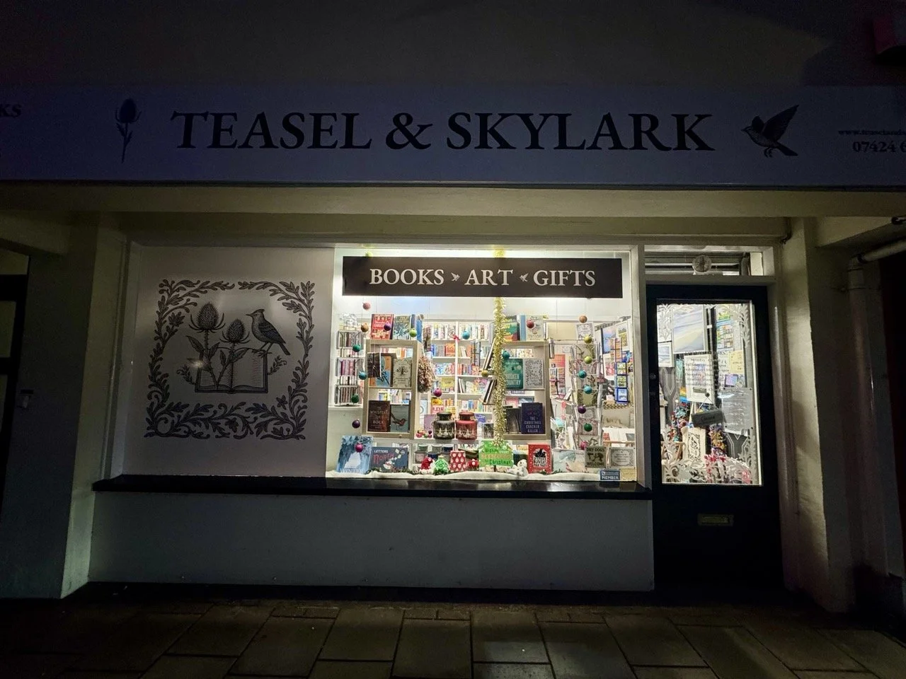Bookstore window display with sign reading 'Books, Art, Gifts'; interior filled with books and decorative items, illuminated from inside.