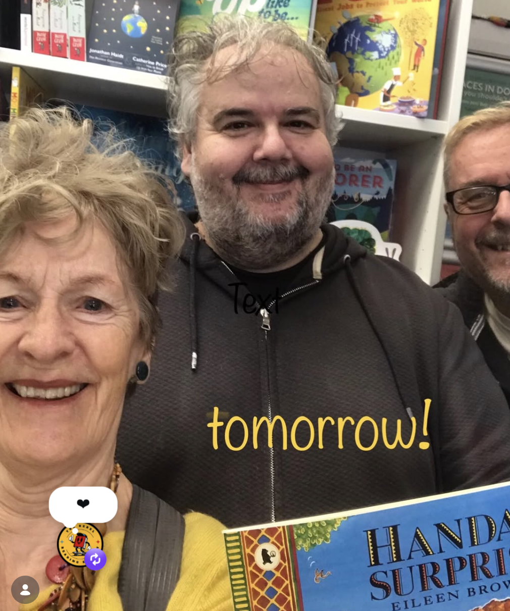 Group of three people taking a selfie inside a bookstore, with bookshelves filled with children's books in the background. The woman on the left has short, curly, light-colored hair, and is smiling. The man in the middle has gray hair and a beard, we