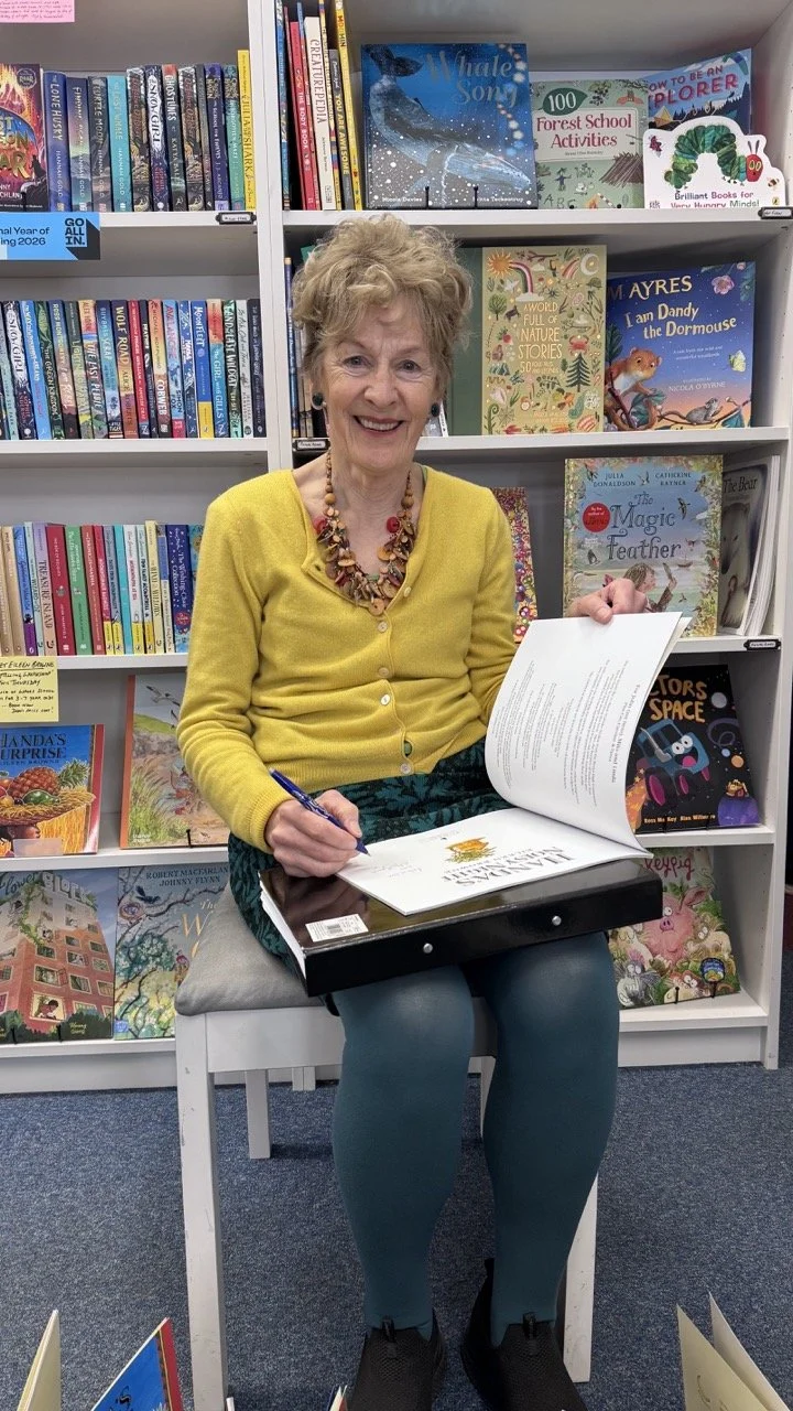 A smiling elderly woman with short, curly blonde hair, wearing a yellow cardigan, a colorful beaded necklace, teal tights, and black shoes, sitting on a white chair in front of a bookshelf filled with children's books in a library. She is holding an 