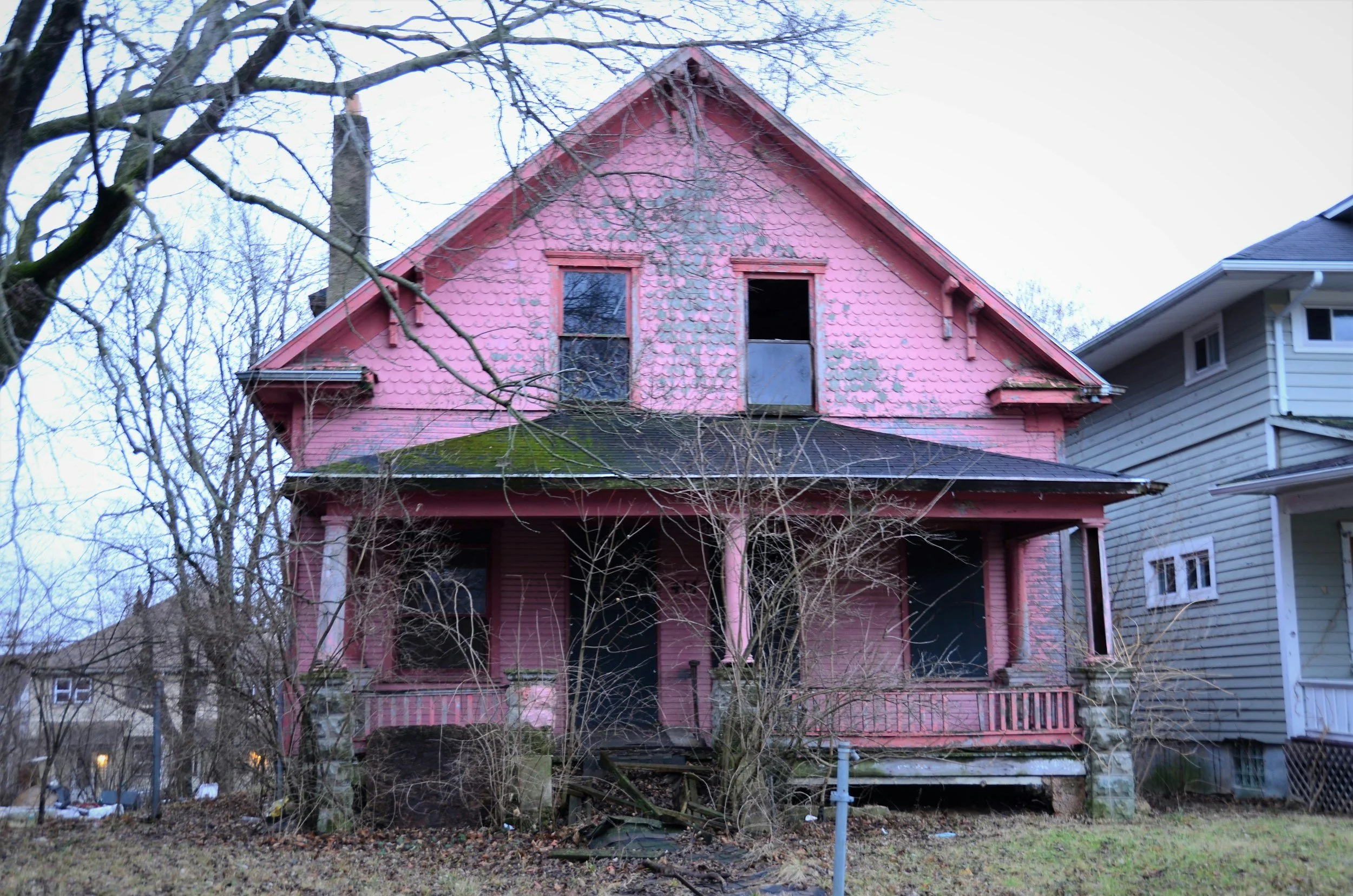Old, abandoned two-story pink house with peeling paint, broken windows, overgrown bushes, and leafless trees in the front yard.