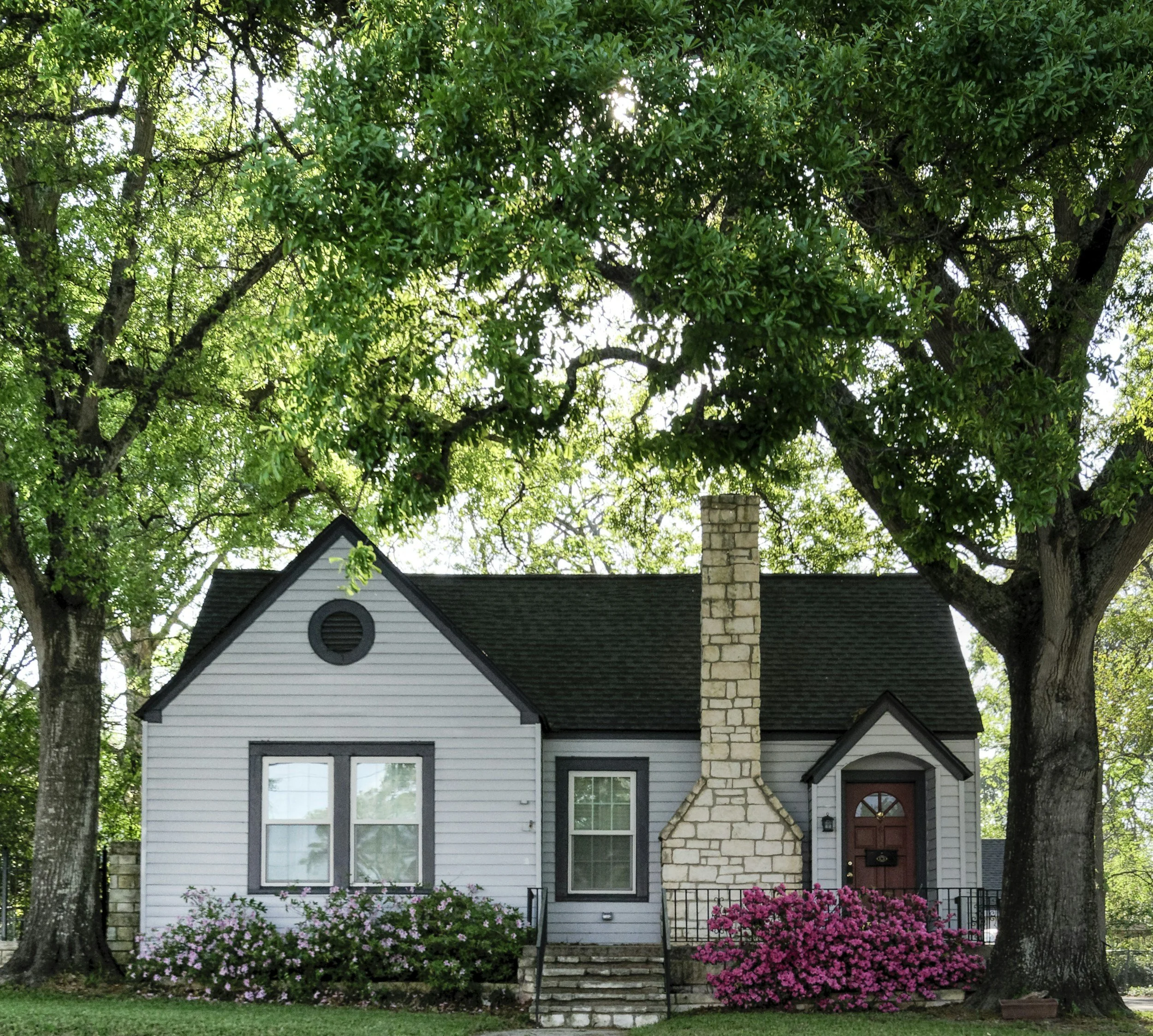 A cozy white house with a stone chimney, surrounded by blooming pink flowers and large leafy trees, with sunlight filtering through the branches.