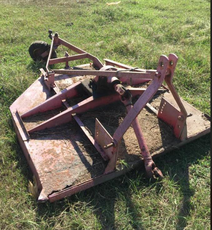 A rusty, pink ATV mower or rotary cutter lying on the grass in a field.