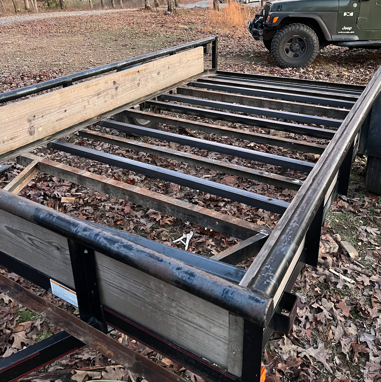 A trailer frame with metal and wooden sides parked outdoors on fallen leaves, with a green Jeep in the background.
