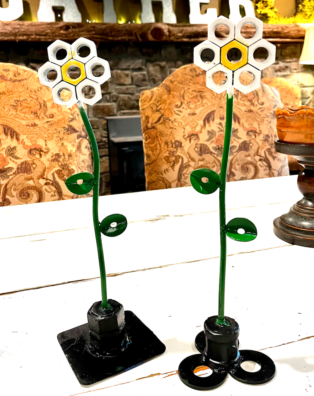 Two decorative metal flowers with hexagonal petal shapes arranged in a flower pattern, mounted on green stems with two leaves each, placed on black bases on a white wooden table.