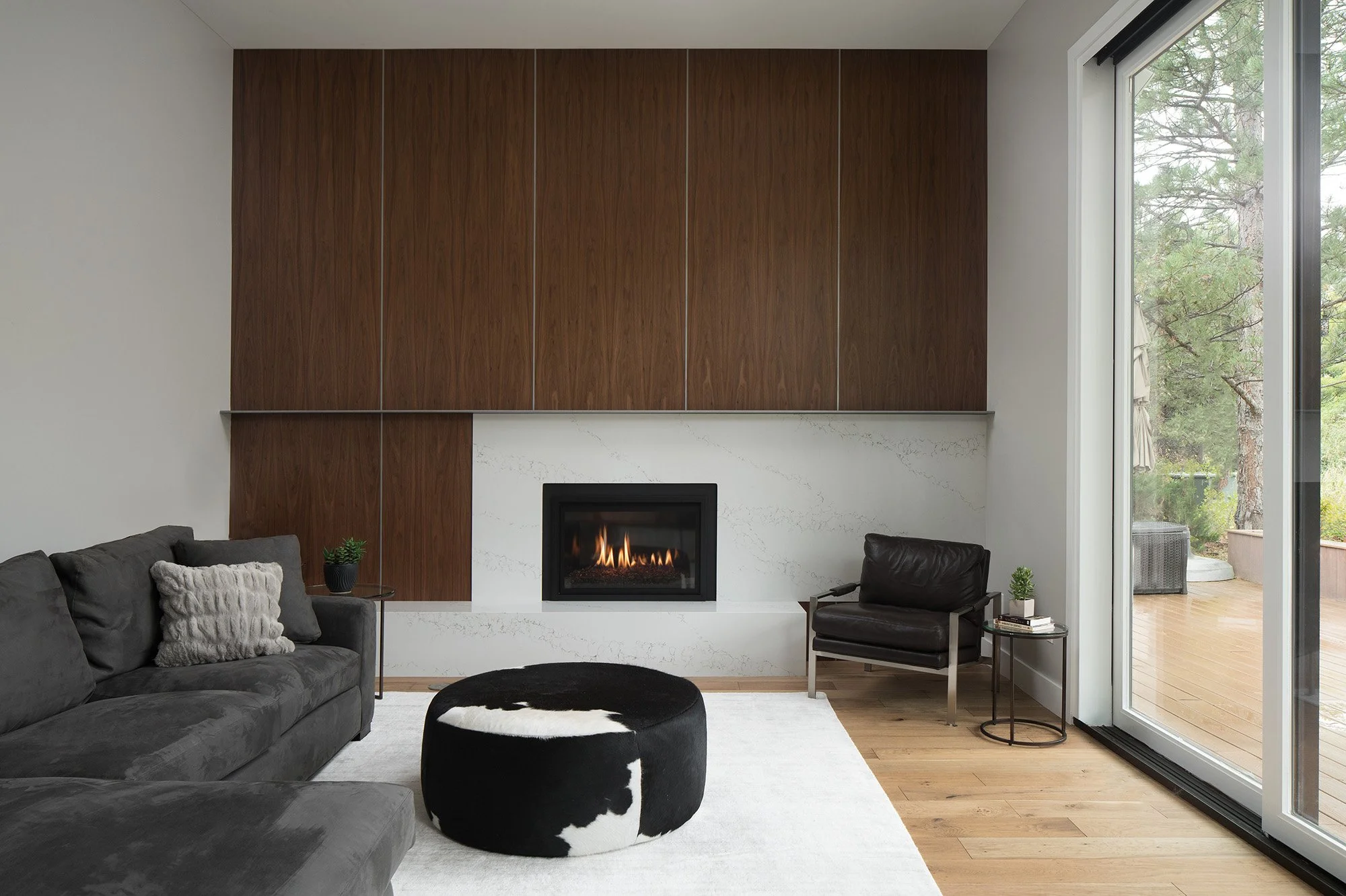Modern living room with a sculpted fireplace wall, walnut paneling, and expansive sliding glass doors opening to the surrounding landscape.