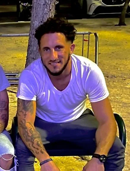 Young man with dark curly hair, beard, and tattoos on his left arm, sitting on a bench outdoors at night, wearing a white T-shirt and dark pants.