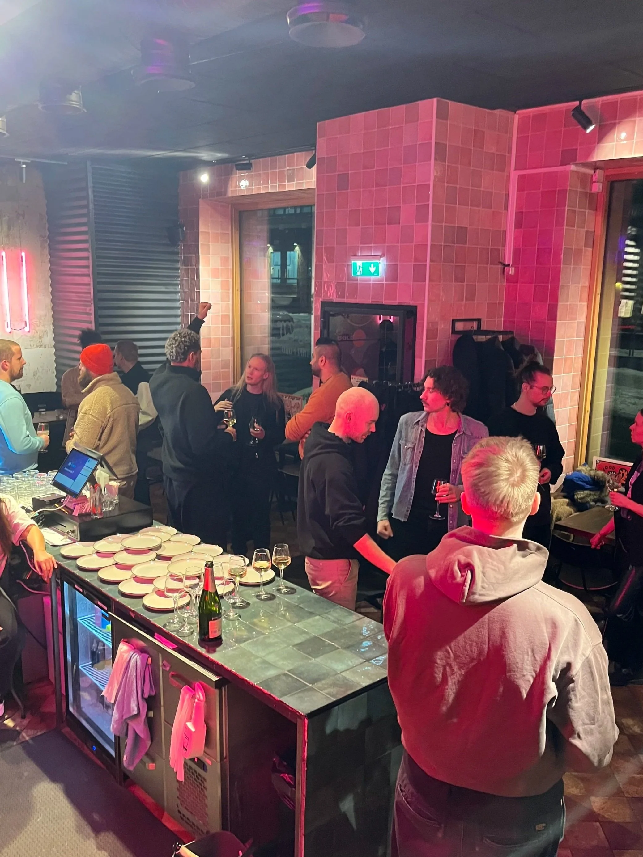 People socializing and drinking at a bar or lounge with pink tiled walls, a table with empty plates and champagne bottles, and a neon sign in the background.