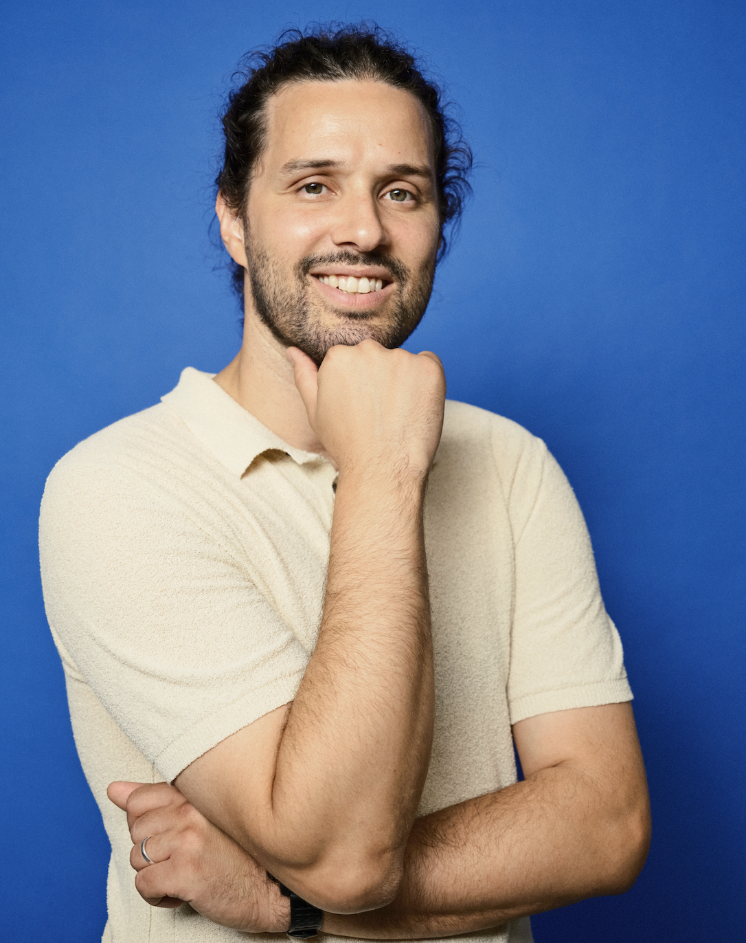 A smiling man with curly hair and rings on his fingers, sitting with clasped hands, against a background of gold streamers.