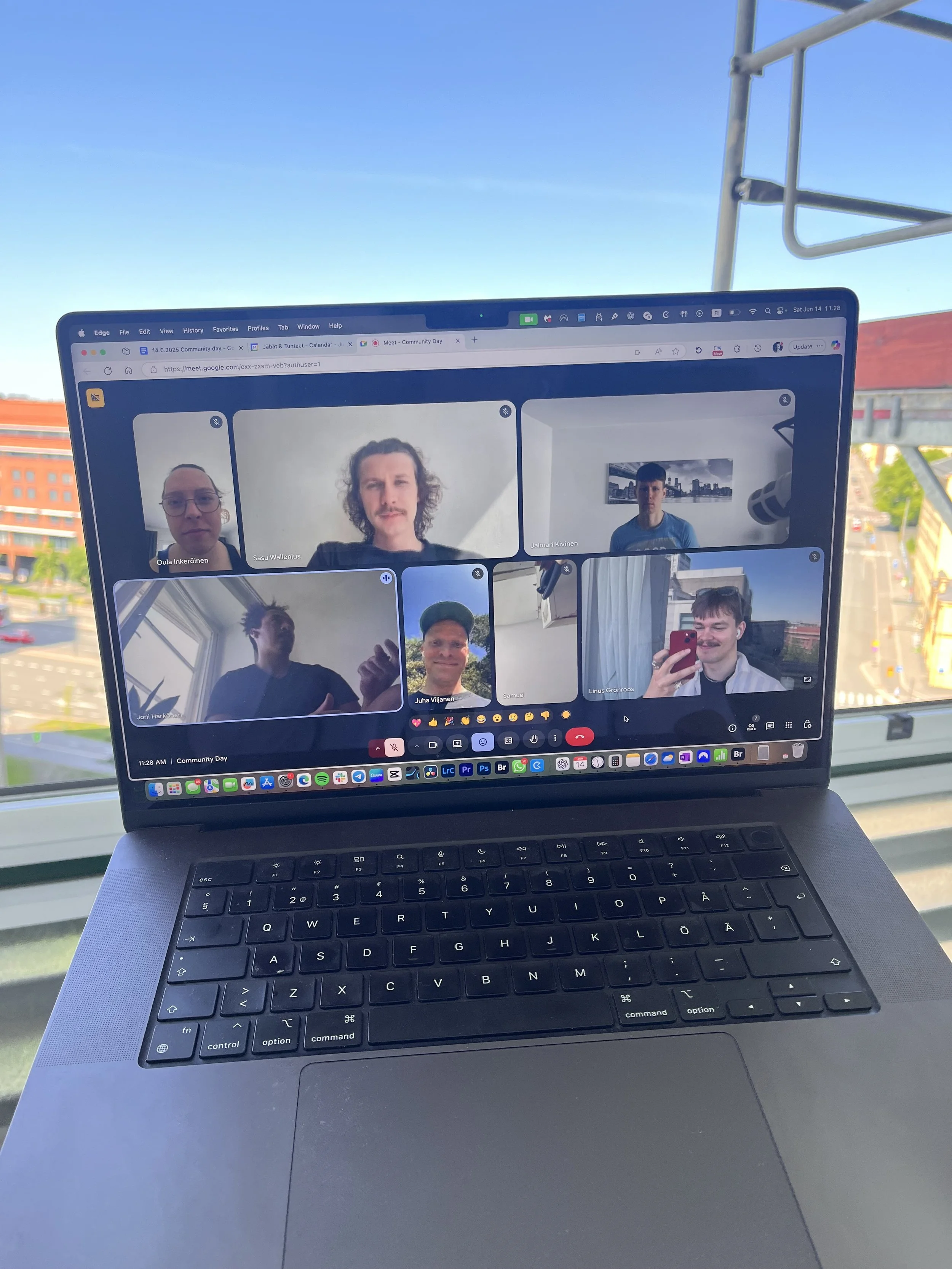 Laptop screen displaying a video call with eight participants, some giving thumbs up, on a desk near a window with a city view behind.