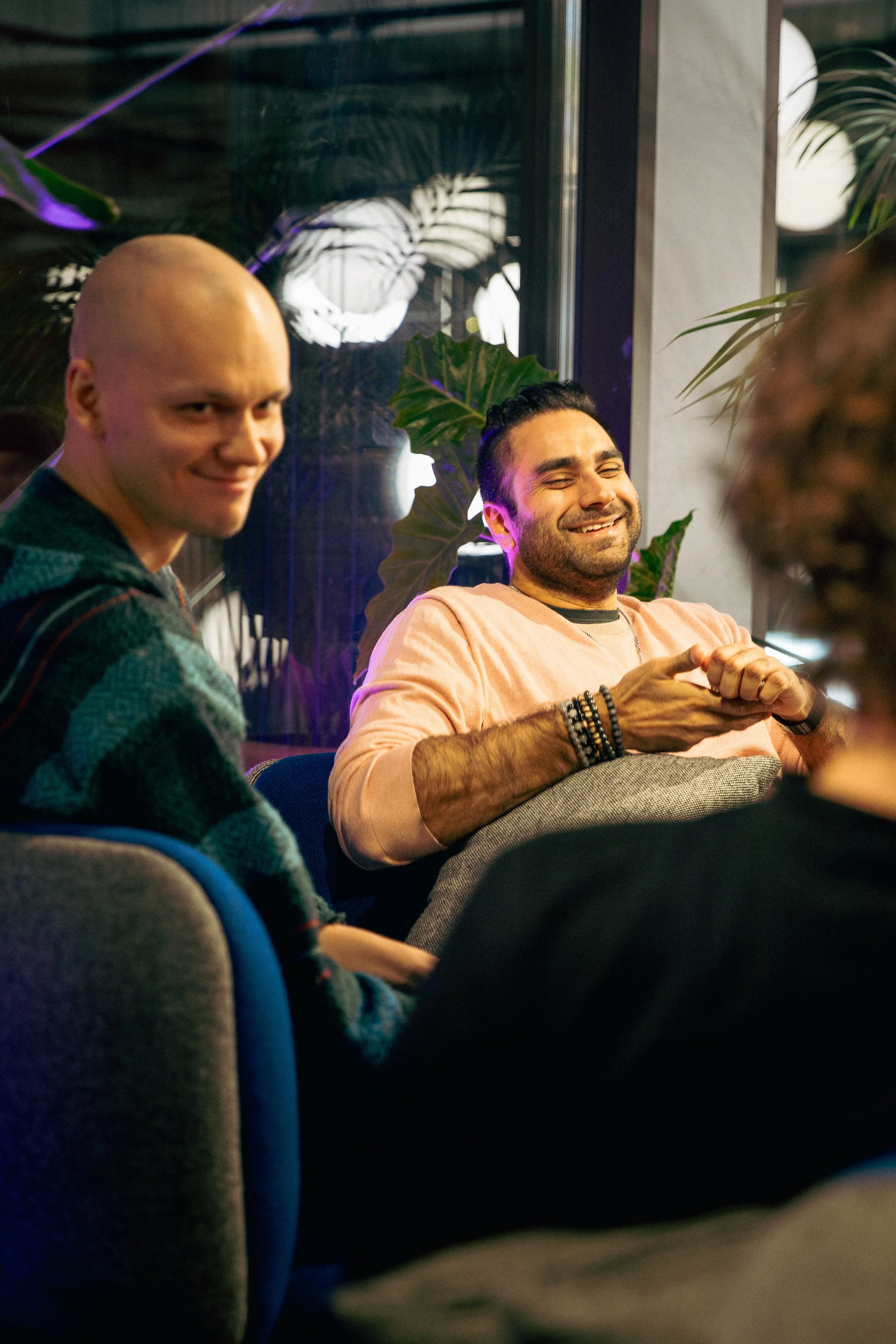 Three men sitting on a couch in a cozy, modern indoor setting with plants behind them. Two of the men are smiling, one with his eyes closed, and the third man is looking towards the camera with a smile.