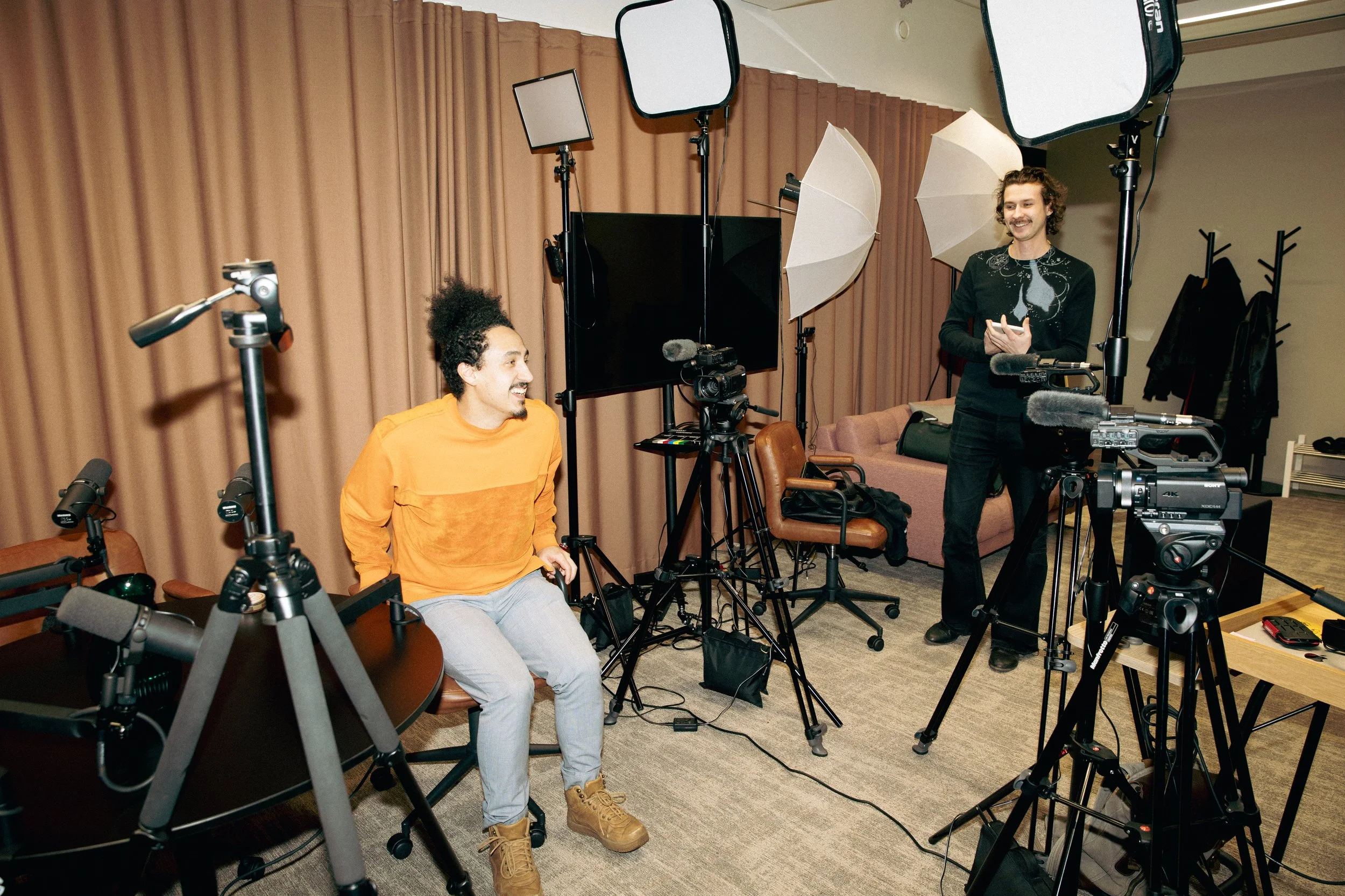 Two men in a photography studio with lighting and camera equipment. One man, seated on a chair, wearing an orange sweater and gray pants, is smiling at the other man, who is standing and holding a device, dressed in black. The studio has beige curtai