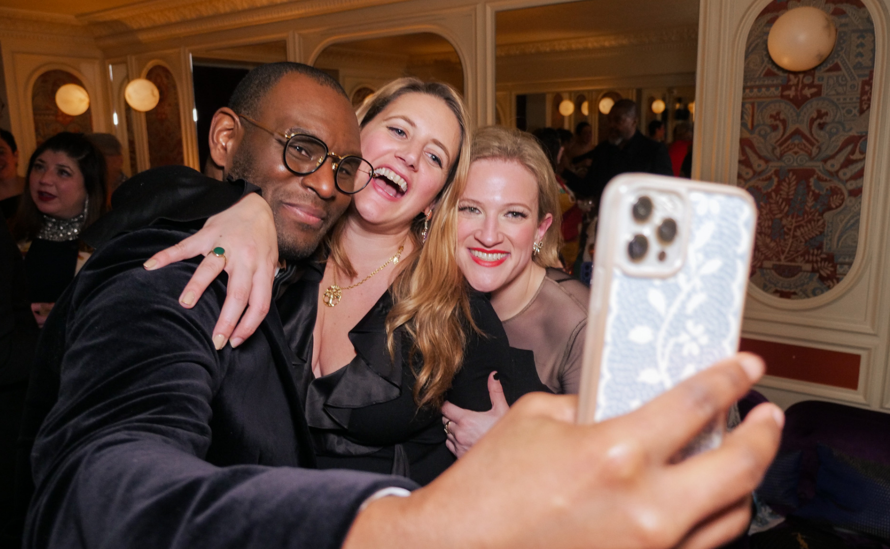 Three friends taking a selfie at a social event, smiling and embracing each other in a decorated indoor setting.