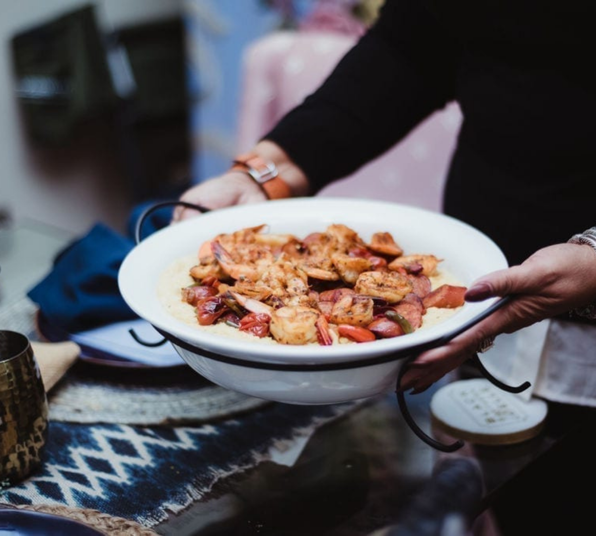 A person holding a white bowl of cooked shrimp and vegetables served over mashed potatoes.