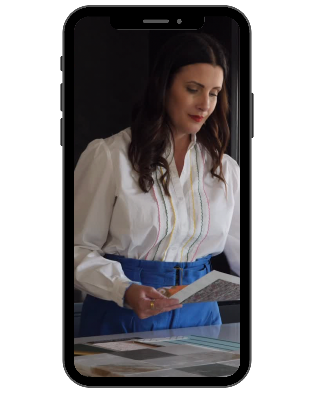 A woman dressed in a white blouse with colorful embroidery and blue pants, standing and reading a newspaper in a well-lit indoor space.