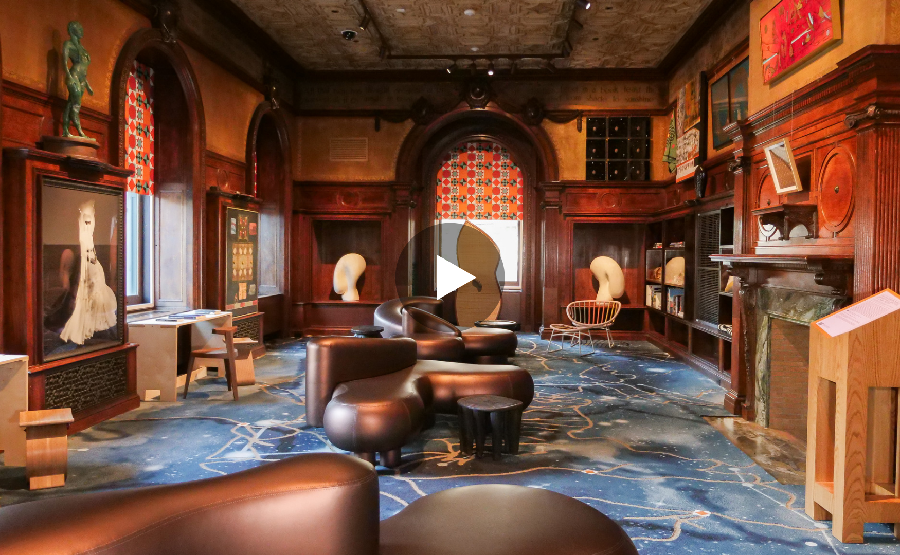 A cozy, art-filled room with dark wood paneling, colorful art on the walls, and eclectic furniture including leather chairs, a small wooden stool, and modern decor elements.