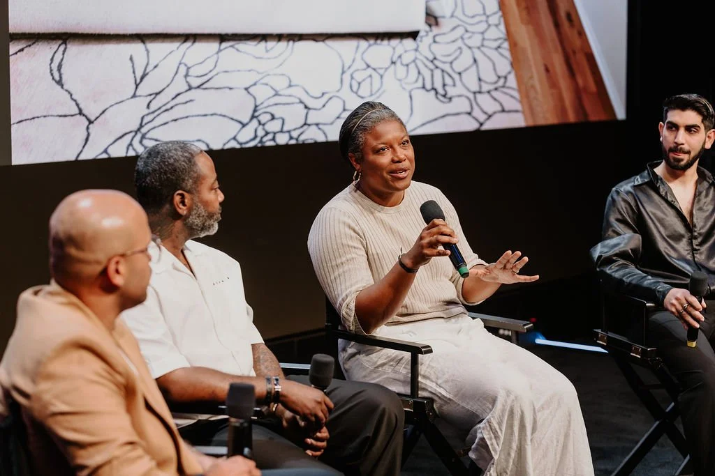 A woman speaking into a microphone during a panel discussion, sitting between four men on stage with a large screen behind displaying an image of a laid carpet with a leaf pattern.