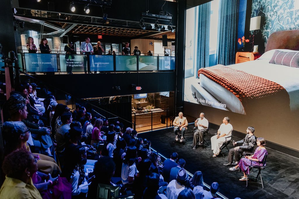 Panel discussion with five people on stage in front of a large audience, a big screen showing a bedroom scene, in an indoor venue with second-floor balcony and audience members seated below.