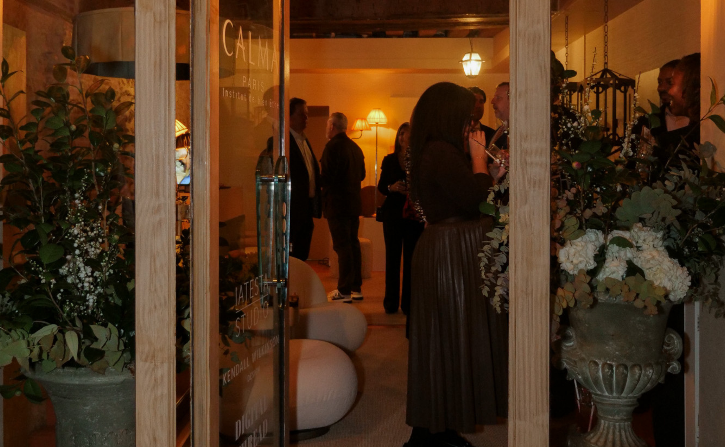 People socializing inside a warmly lit room with floral arrangements and vintage decor, seen through a glass door.