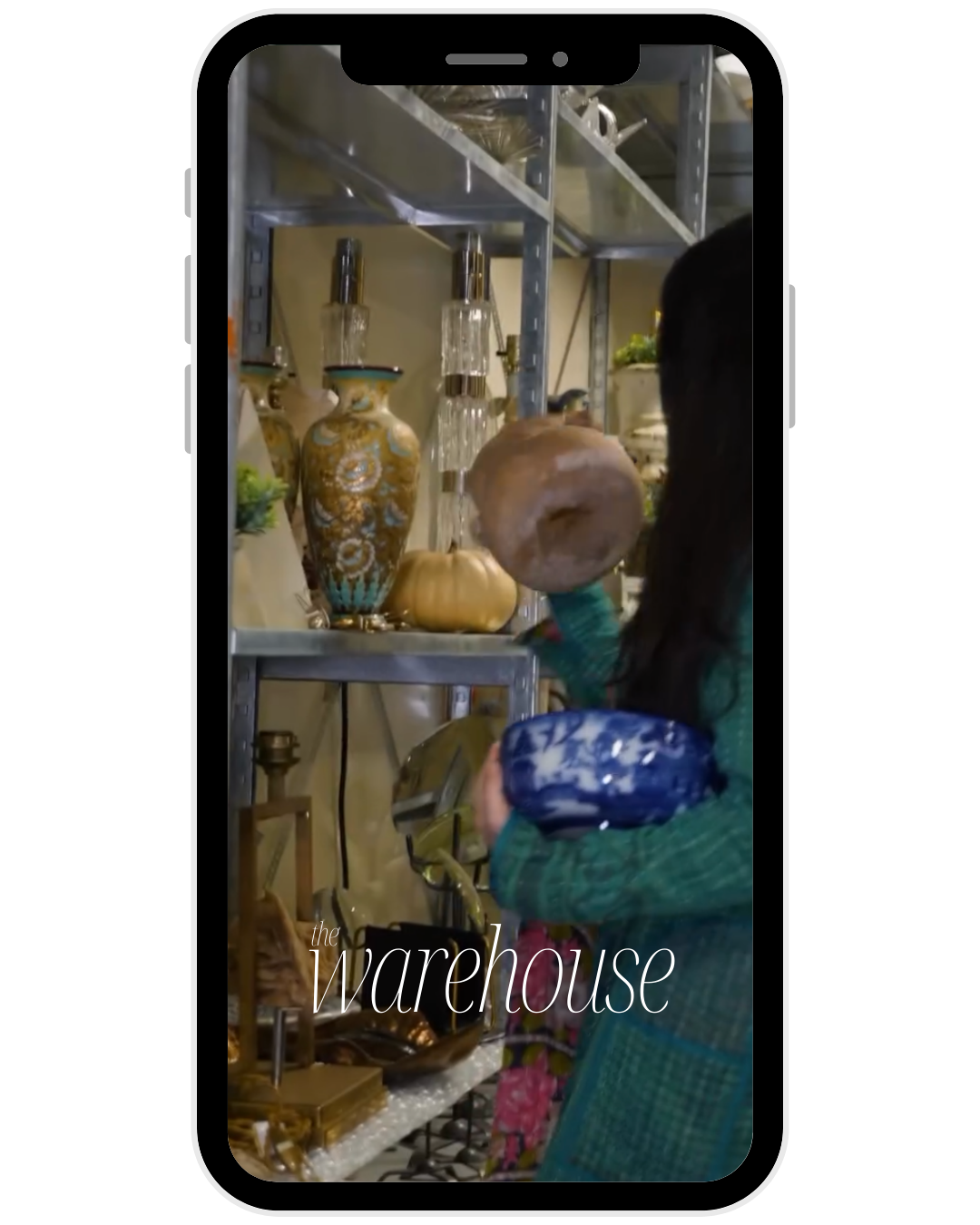 A woman with long dark hair, wearing a green top, is shopping in a warehouse or store, holding a ceramic object. Shelves behind her display decorative vases, gold pumpkins, and windmill lamps.