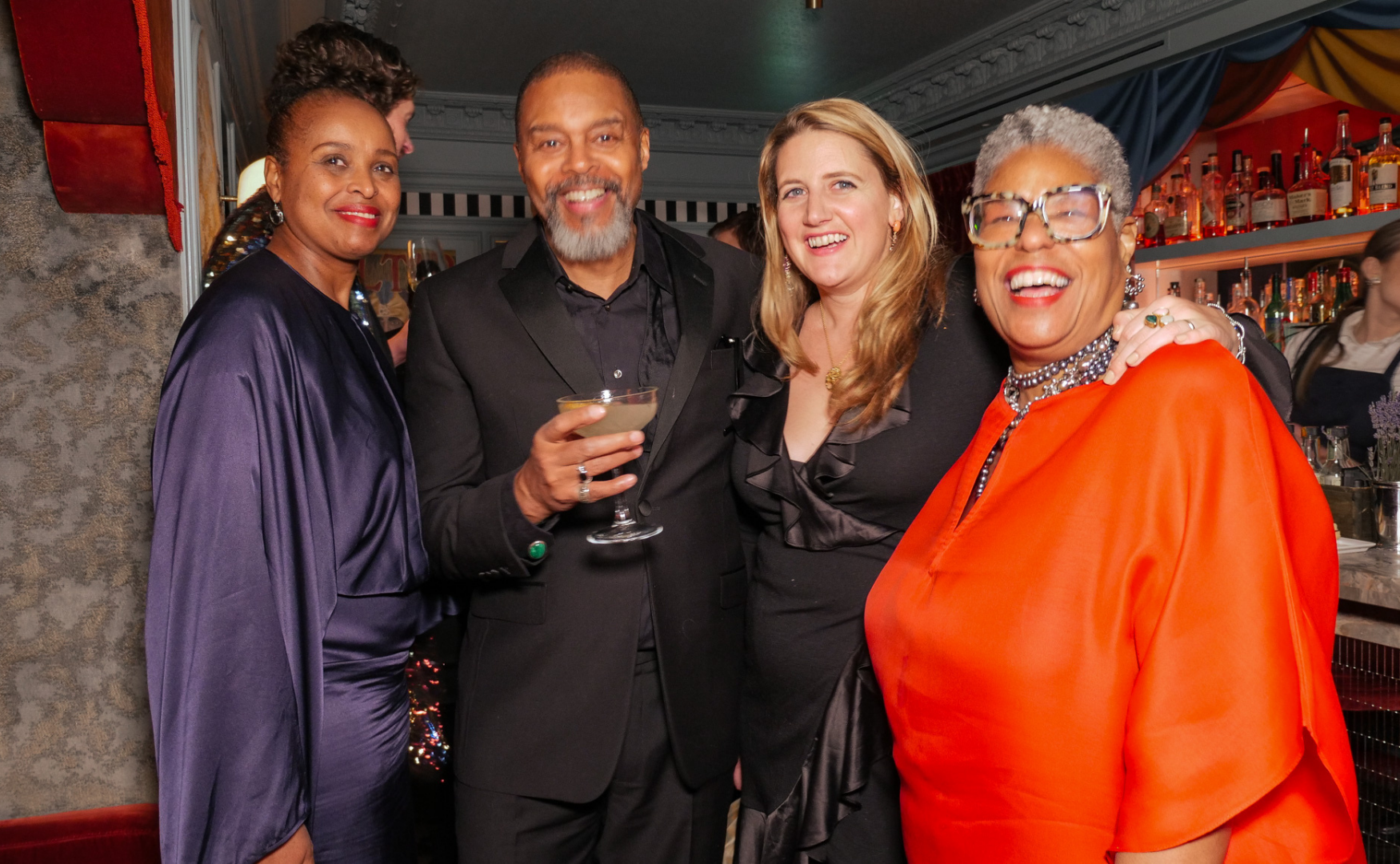 Group of four people dressed in formal attire, smiling at a social gathering, with a bar in the background.