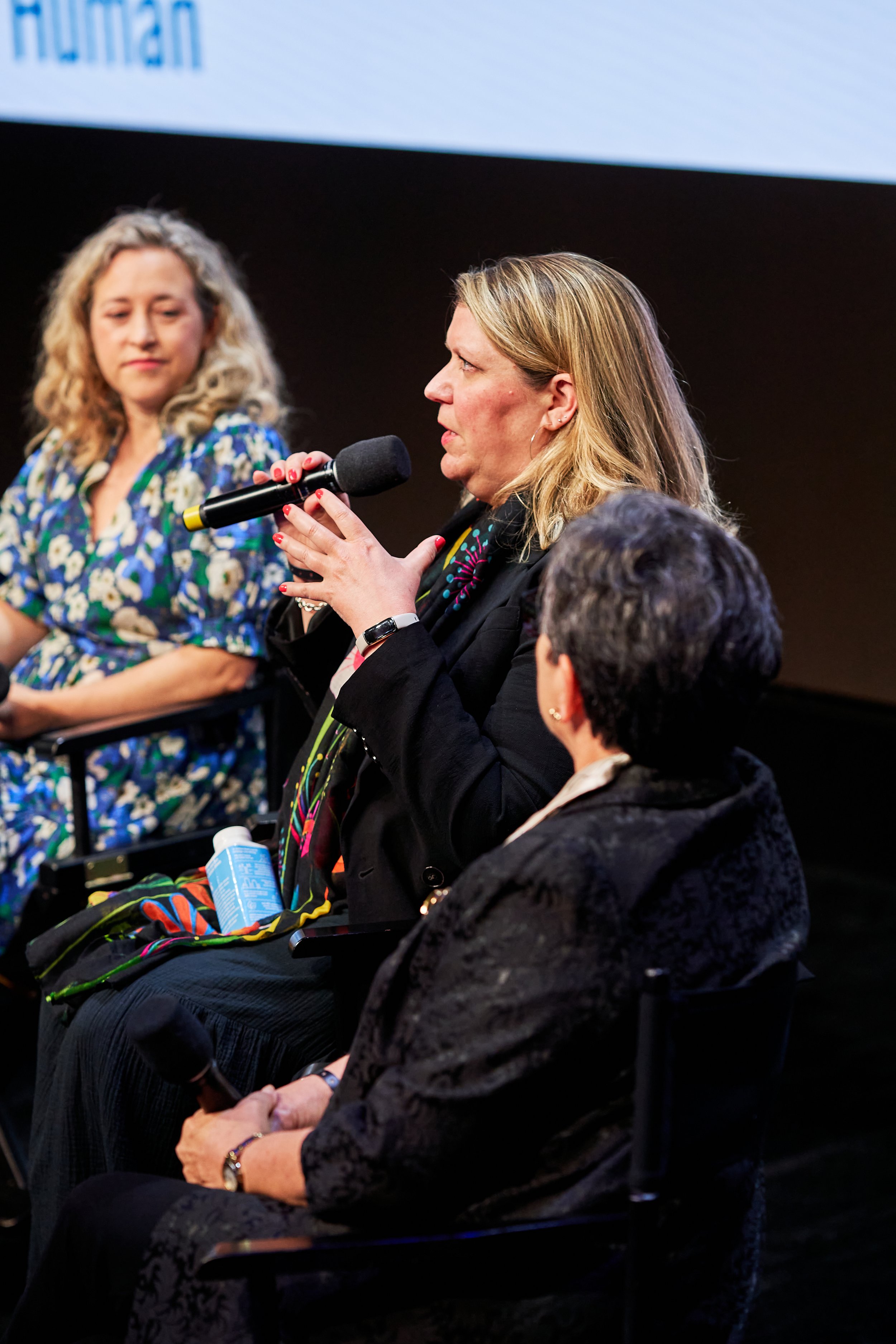 A woman with blonde hair speaking into a microphone while seated on a panel during an event, with two other women listening; one with curly blonde hair in a floral dress, and another with short dark hair in a black jacket.