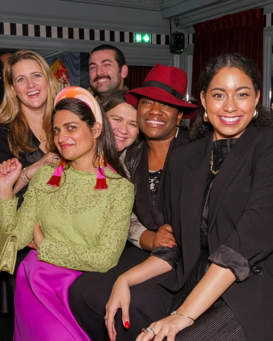Six people smiling and posing closely together at a social gathering; three women in the front and three in the back, with an indoor setting and festive decorations.